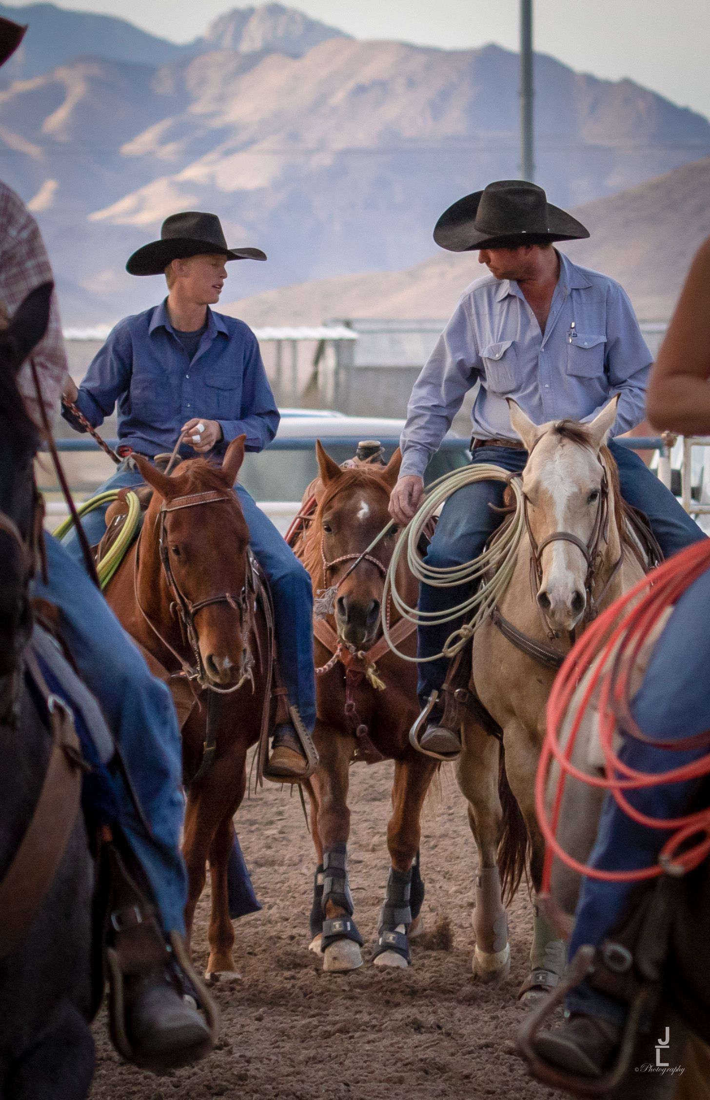 Two cowboys on horseback holding lasso ropes chat together at a dusty rodeo arena at dusk.