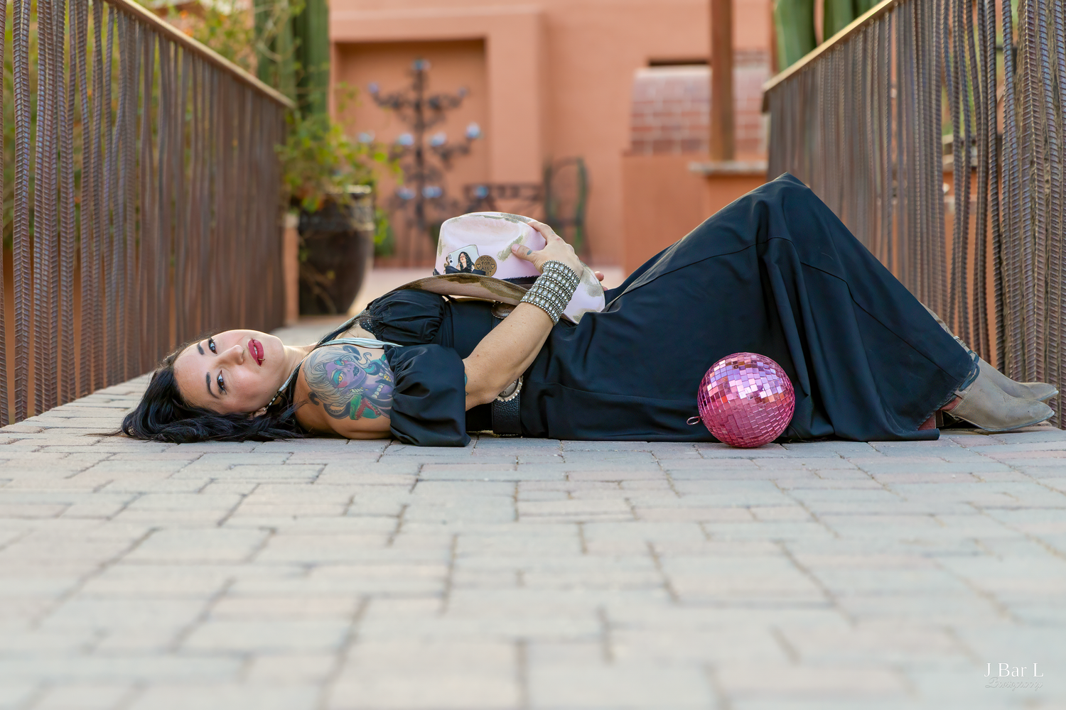 Tattooed woman in black outfit lies on brick pathway with pink disco ball at outdoor urban location.