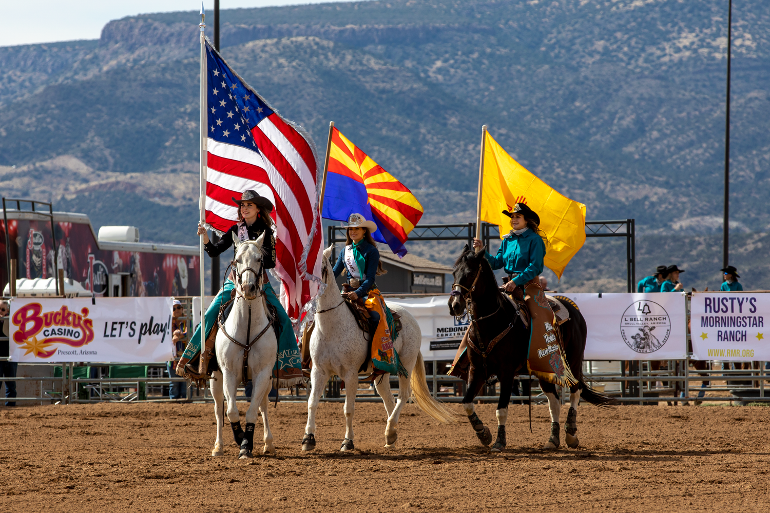 Three riders on horseback carry American, Arizona, and gold flags in a rodeo arena opening ceremony.