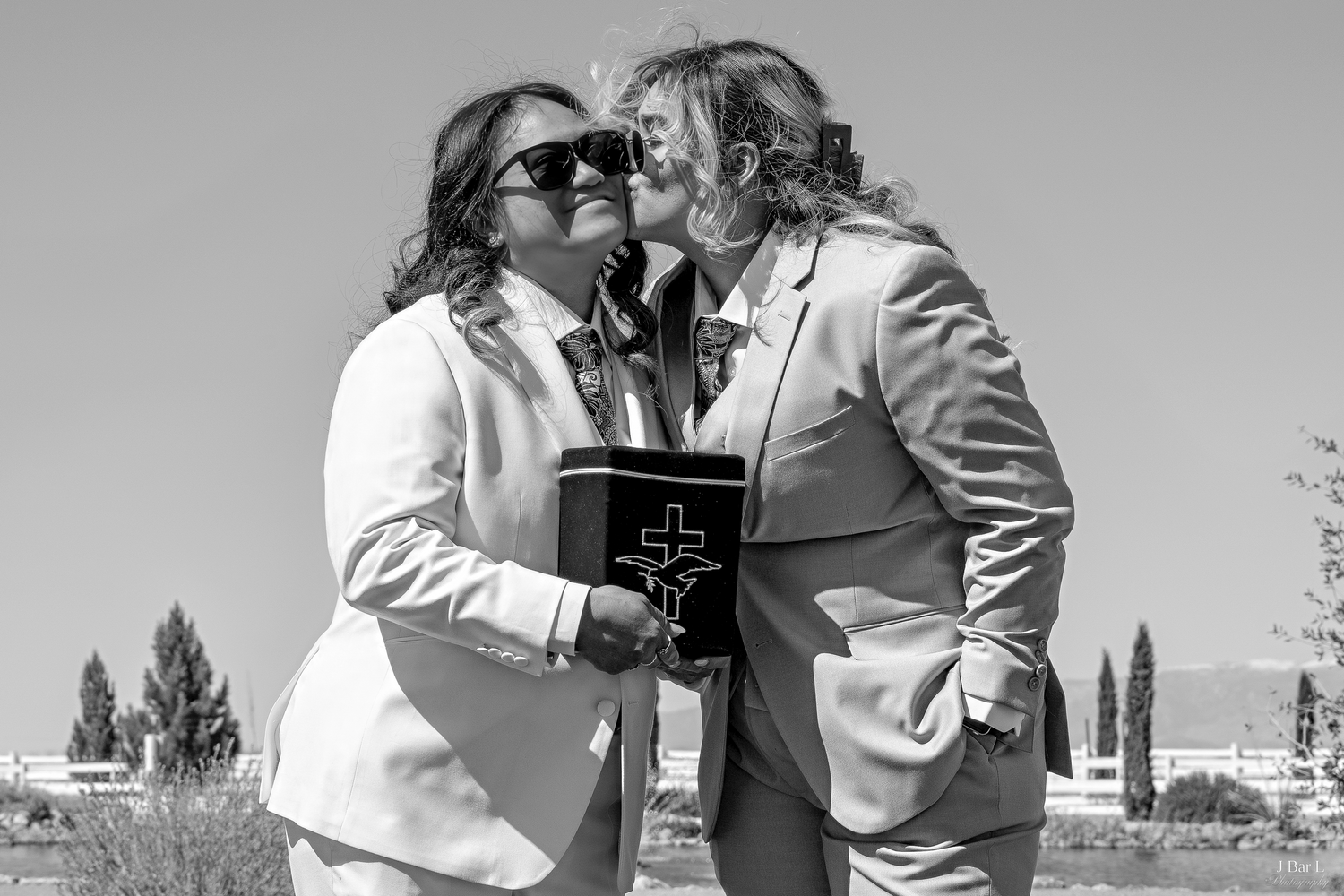 Black and white photo of two brides kissing while holding a bible at an outdoor wedding.