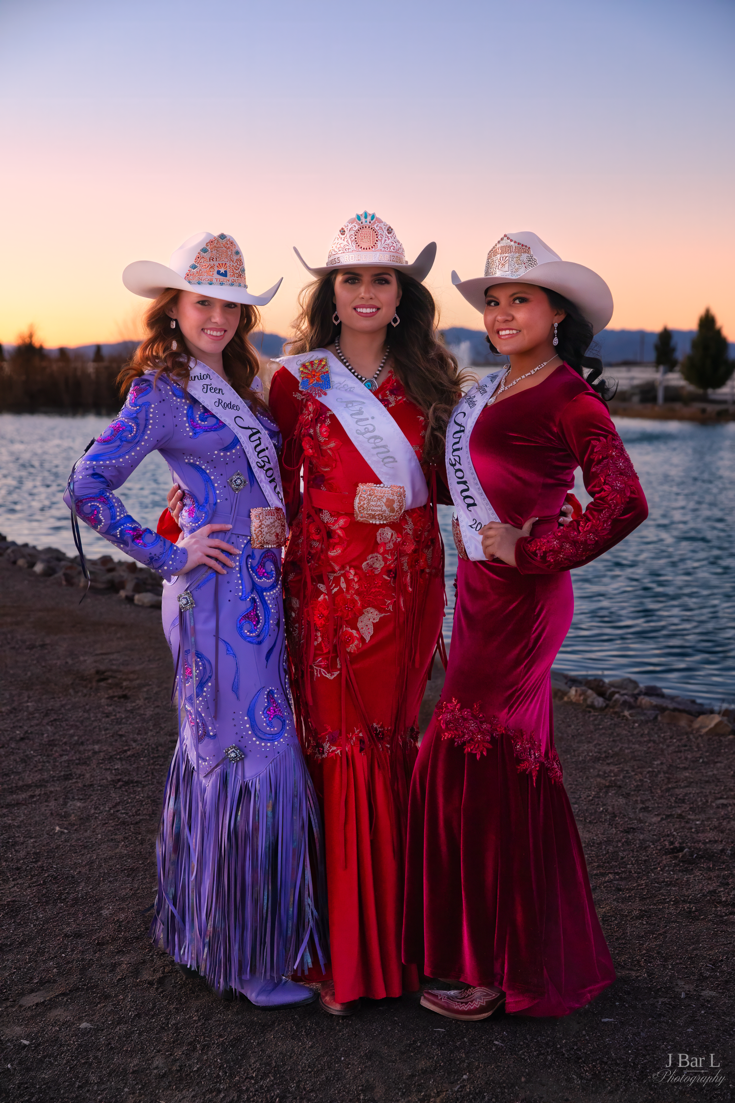 Three women in colorful western pageant gowns and cowboy hats posing at lakeside during sunset.