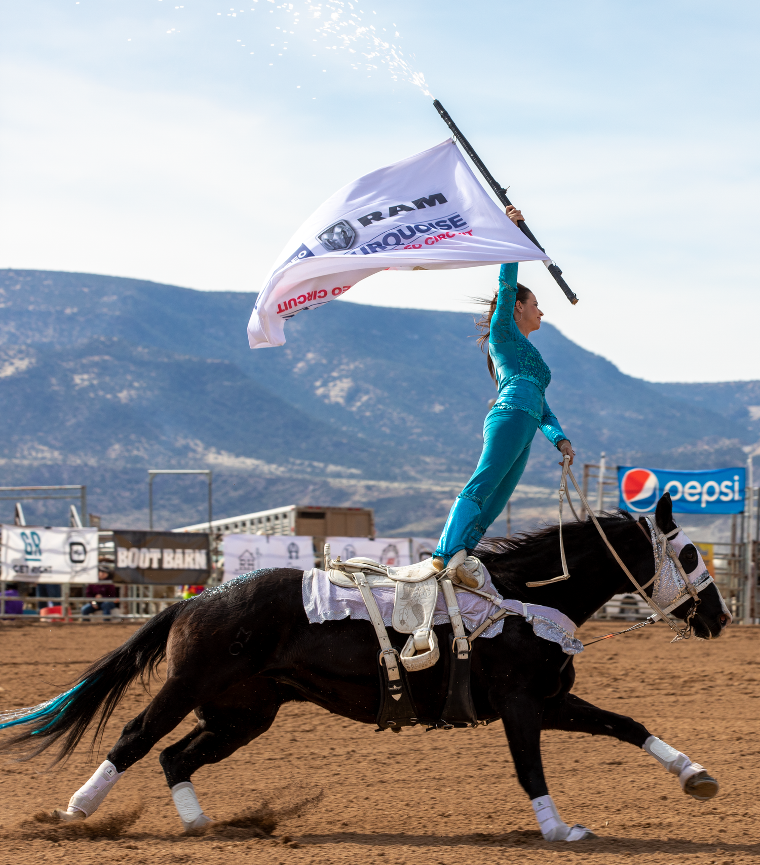 Equestrian performer in teal outfit rides black horse while waving a RAM rodeo sponsor flag.