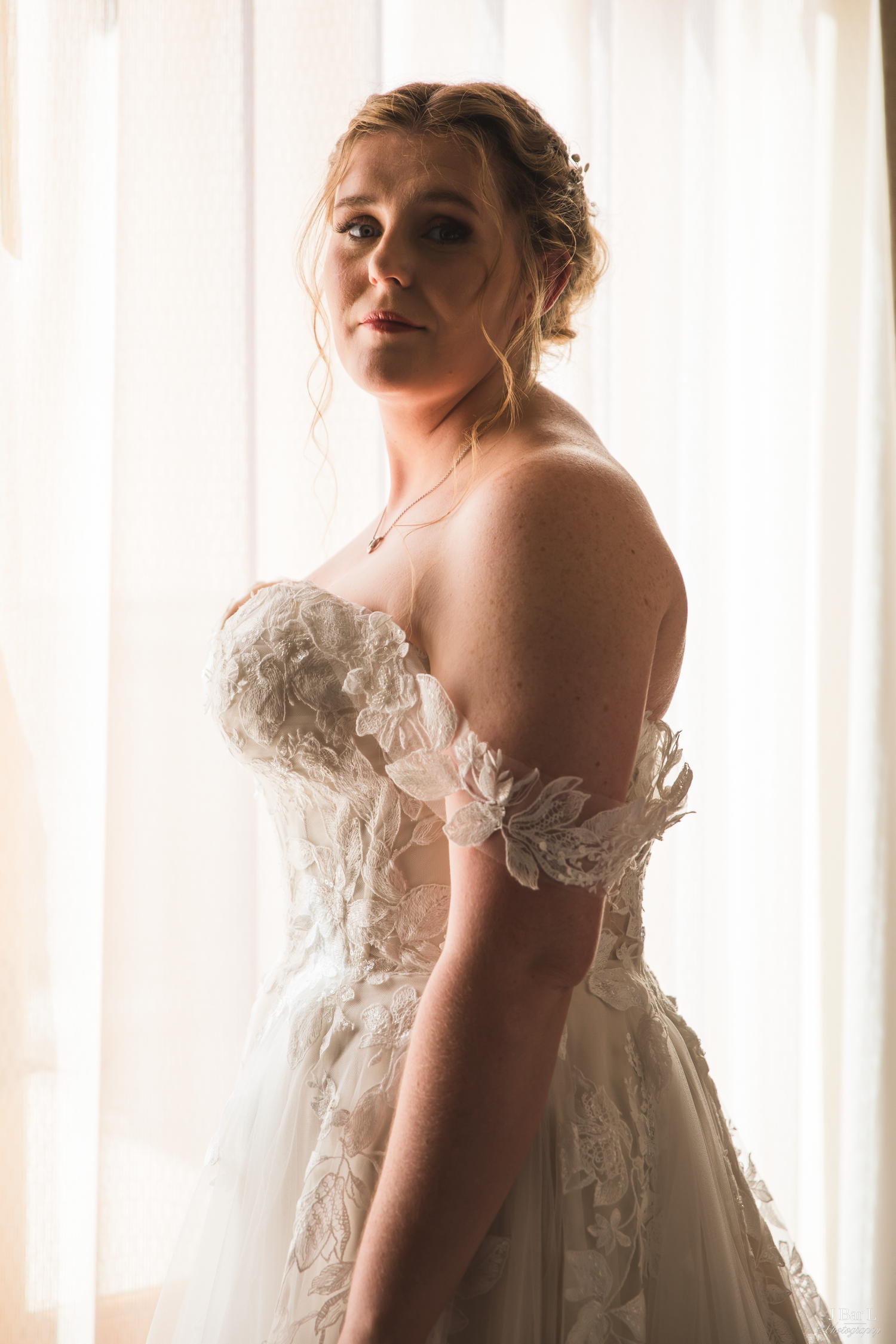 Elegant bride in off-shoulder lace wedding gown posing by a sunlit window.