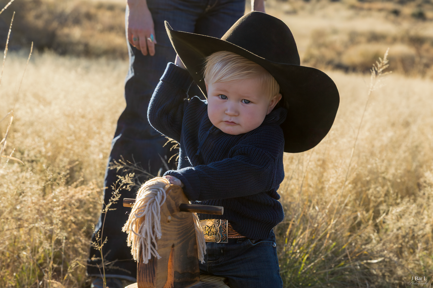 Adorable blonde toddler in a black cowboy hat riding a wooden hobby horse in a golden field.