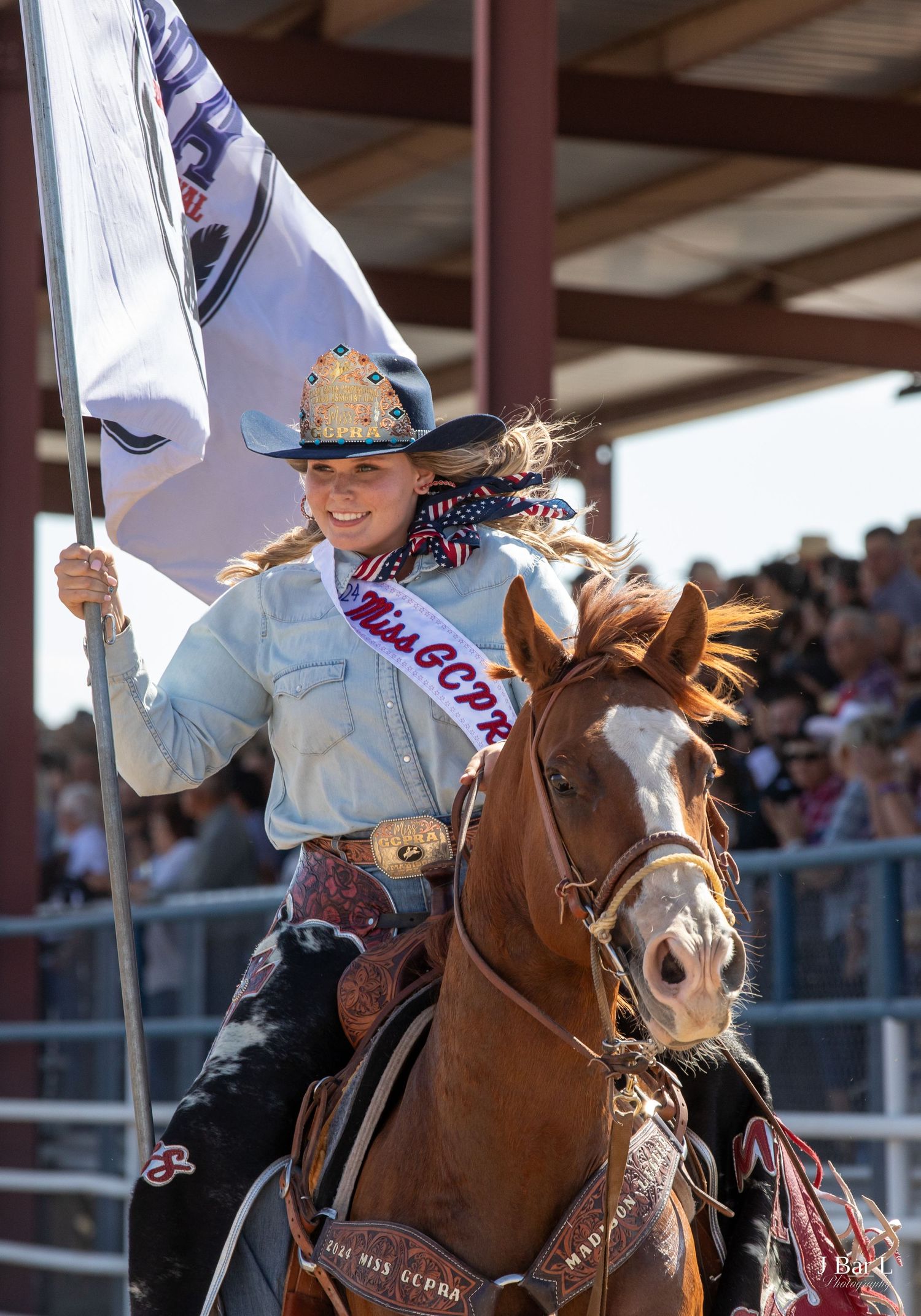 Miss GCPRC rodeo queen on chestnut horse carrying a white flag at a crowded rodeo arena.