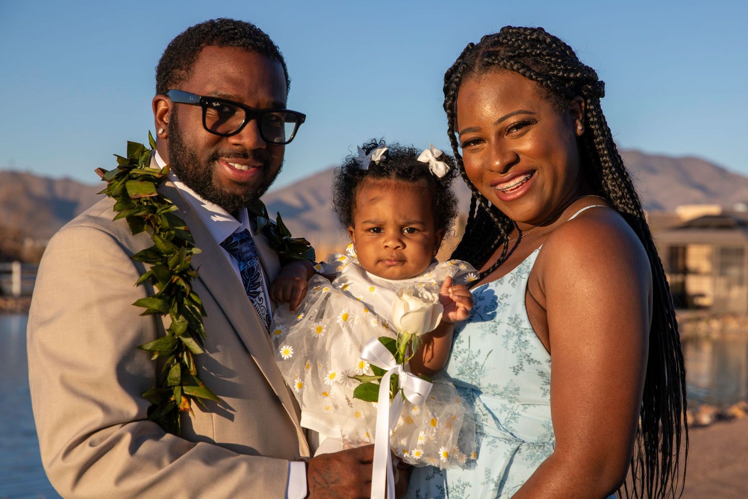 Happy family of three poses at golden hour waterfront, father wearing Hawaiian lei holding baby girl.