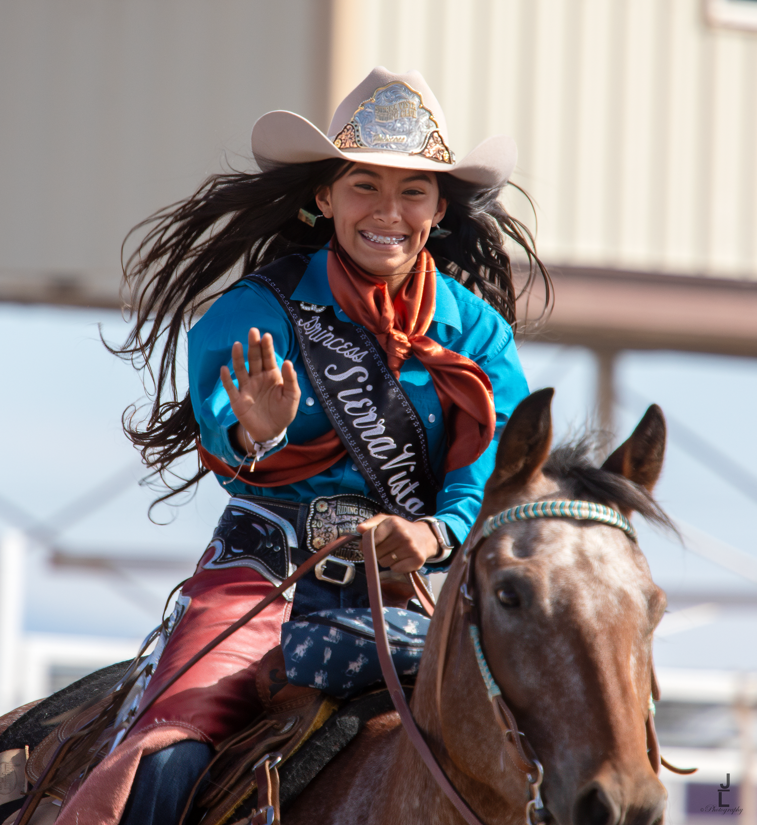 Smiling rodeo queen in blue shirt and sash waves while riding a horse at a rodeo event.