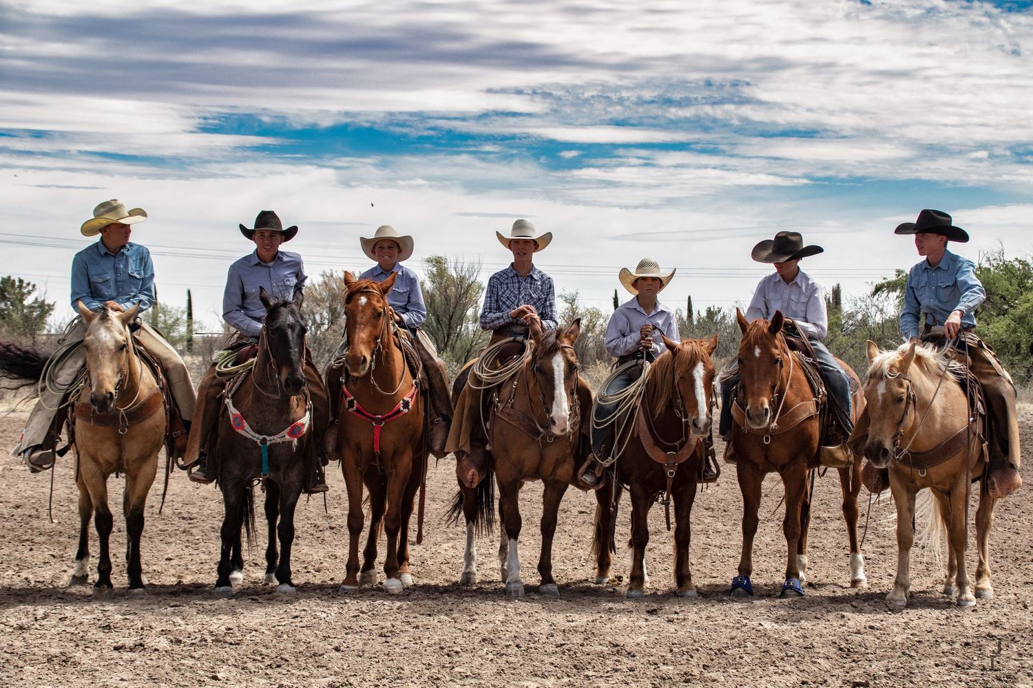 Seven cowboys on horseback lined up in a row wearing cowboy hats in an arid Western landscape.