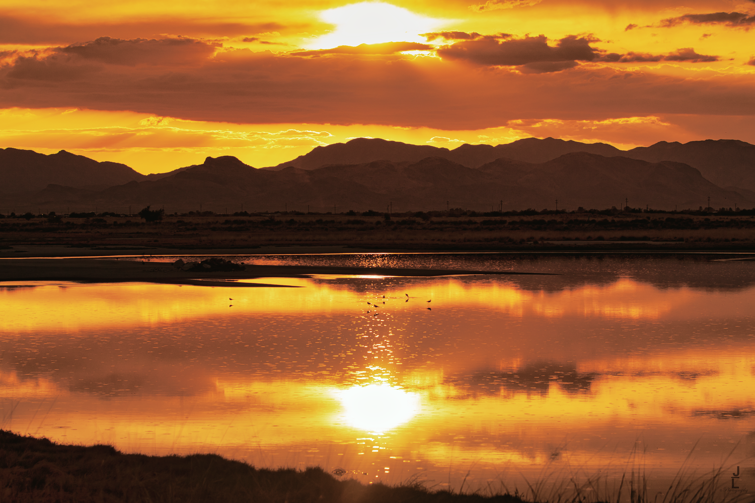 Vivid orange sunset reflecting over a calm lake with mountain silhouettes on the horizon.