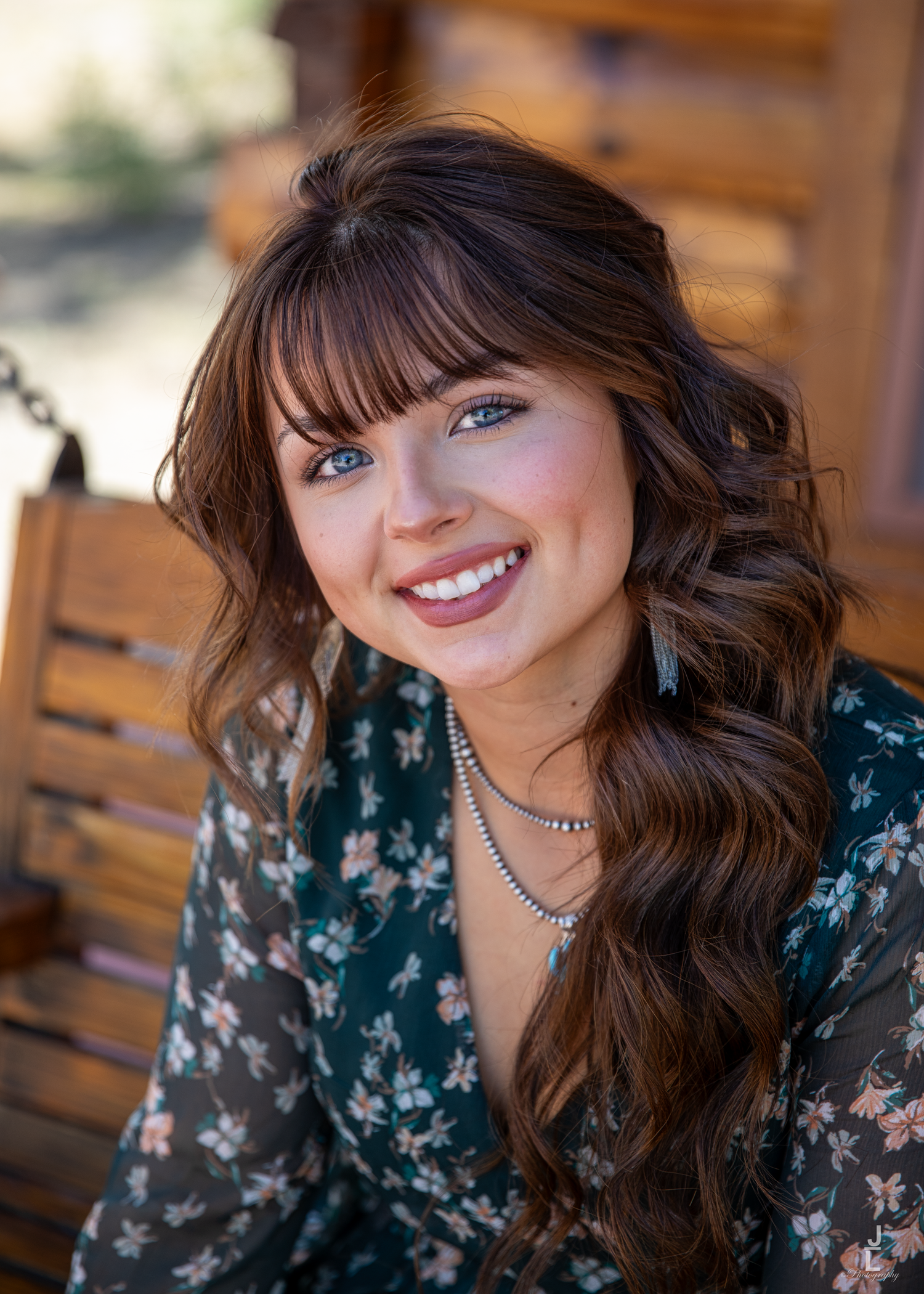 Young woman with brown wavy hair and blue eyes smiling on a wooden swing wearing a floral dress.