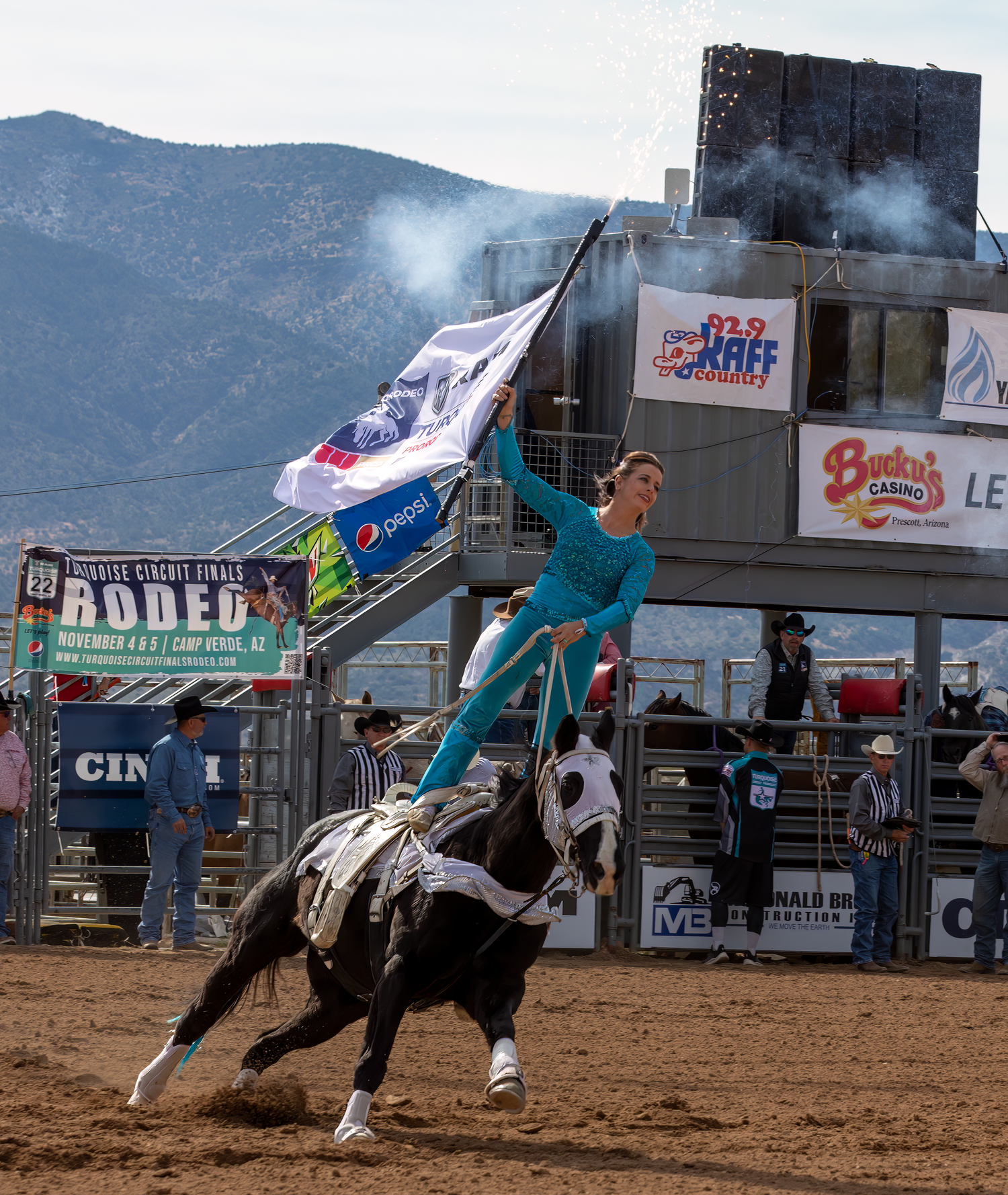 Performer in teal outfit stands on galloping horse waving flag at an outdoor rodeo arena.