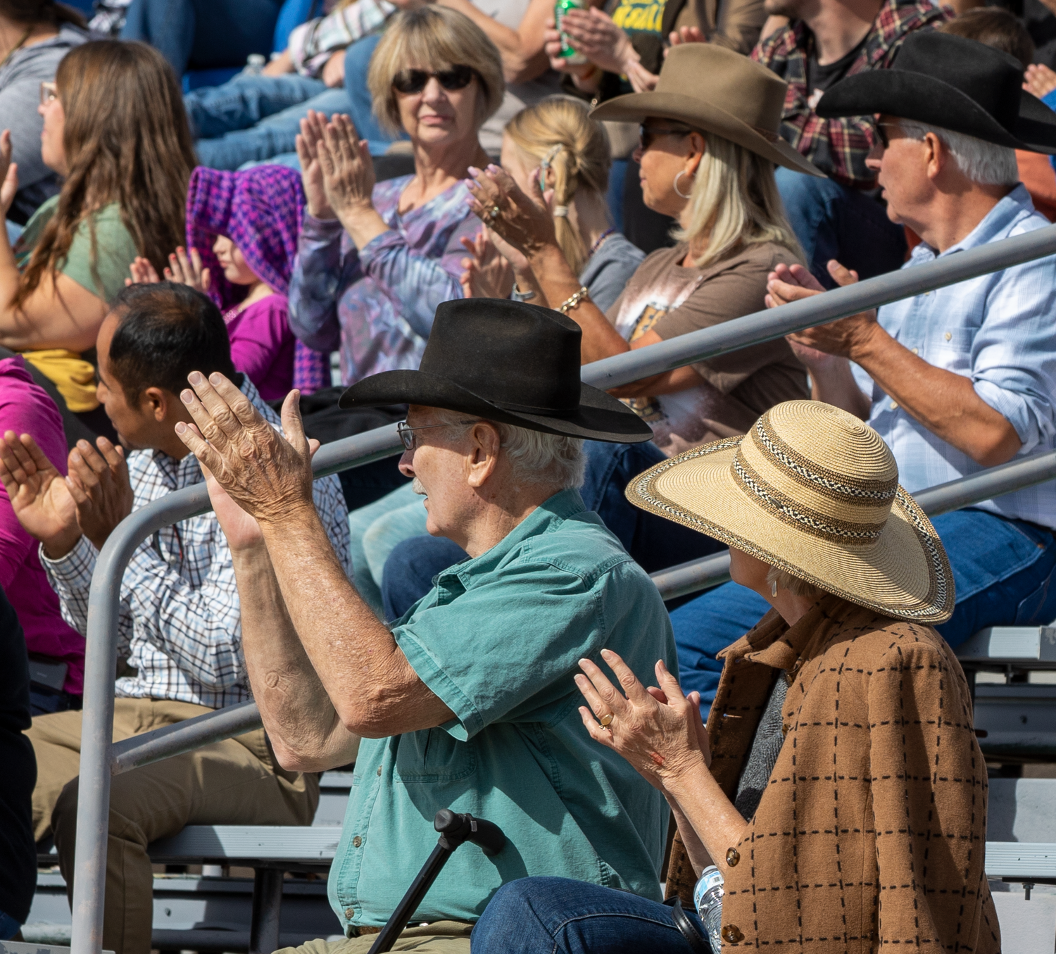 Rodeo crowd of spectators in cowboy hats clapping and cheering enthusiastically in outdoor bleachers.