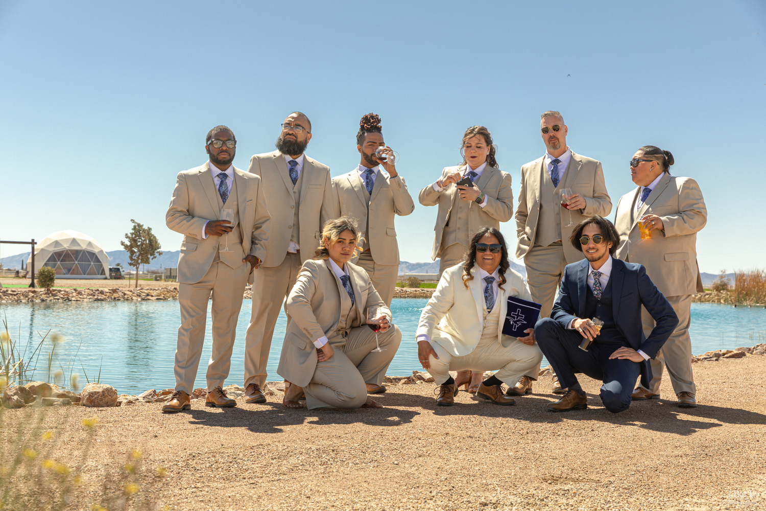 Group of eight men in tan suits pose near a lake on a bright sunny day.