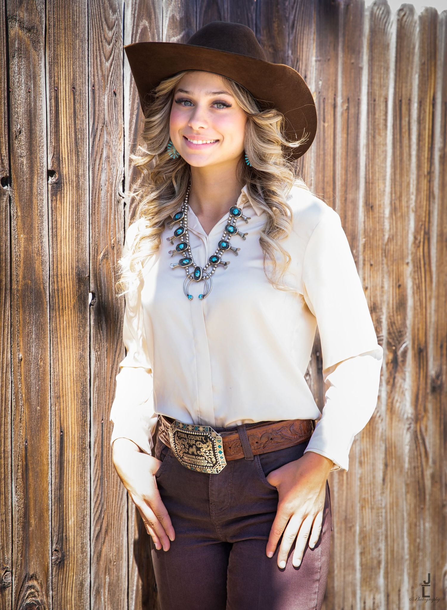 Blonde woman in cream western shirt with turquoise squash blossom necklace and brown hat by wood fence.