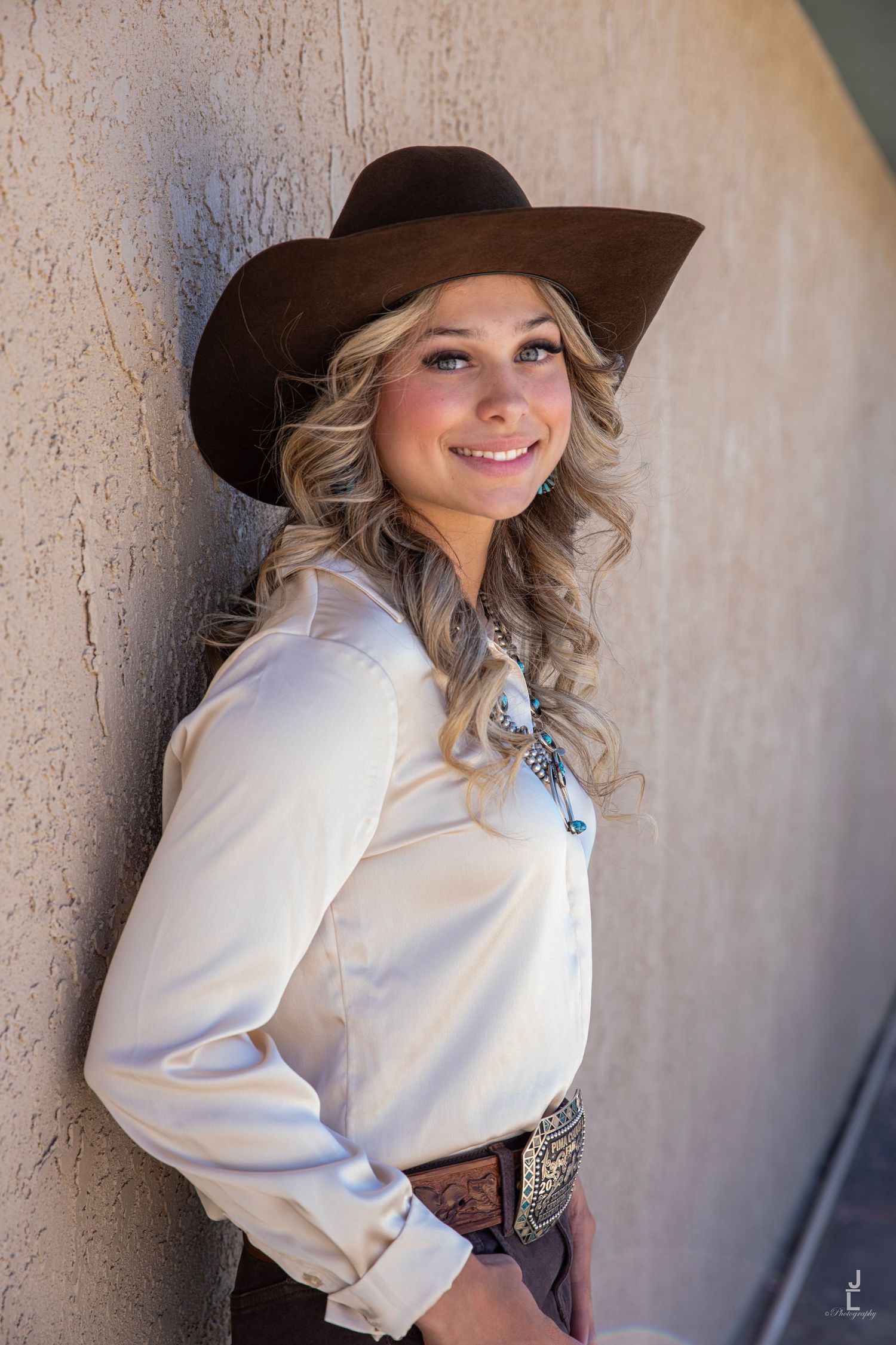 Smiling blonde woman in white western shirt and brown cowboy hat leaning against a tan wall.