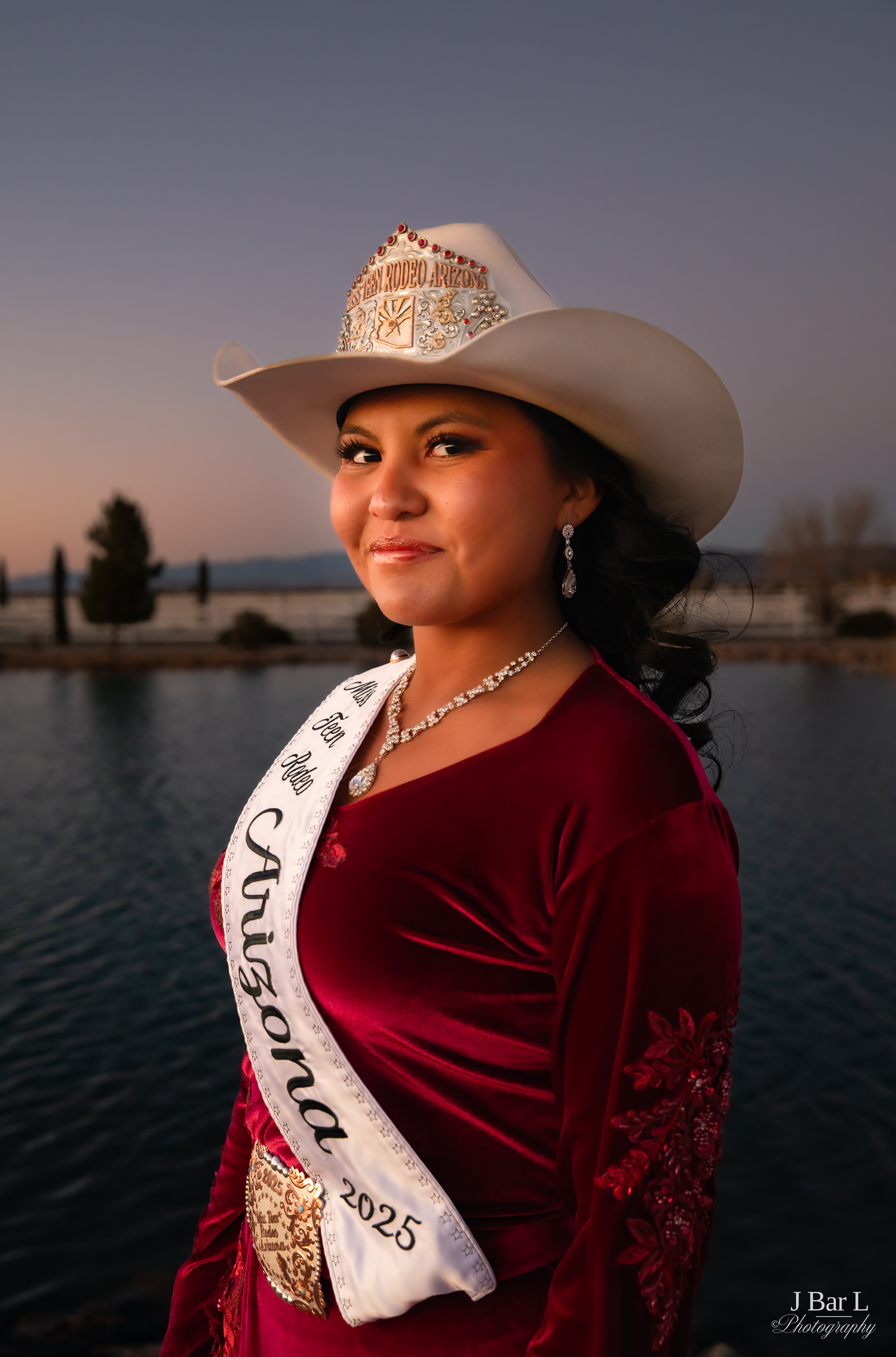 Young woman wearing Arizona 2025 rodeo queen sash and white cowboy hat smiling at sunset.