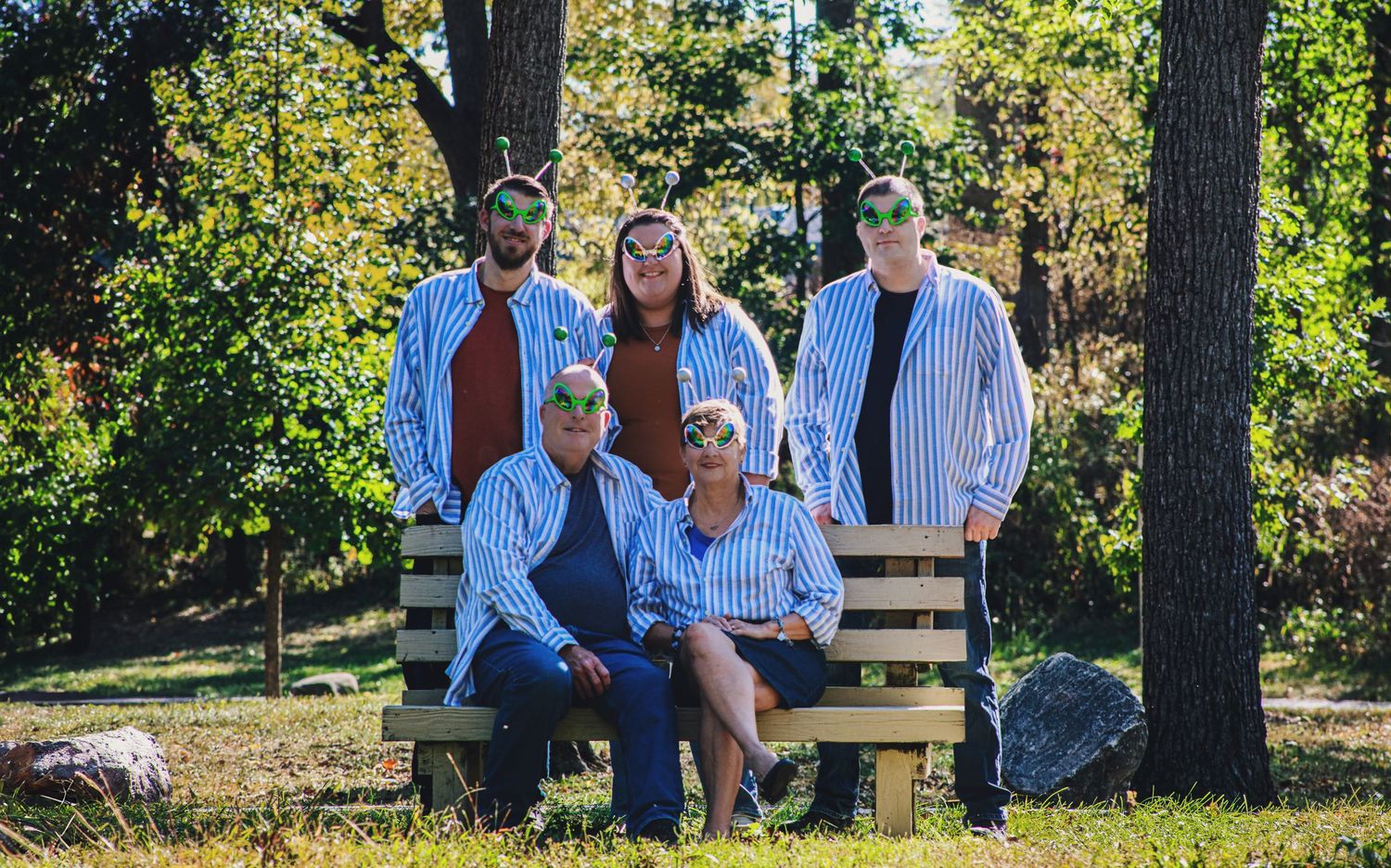 Family of five in matching striped shirts posing together on a wooden bench in a sunny park.