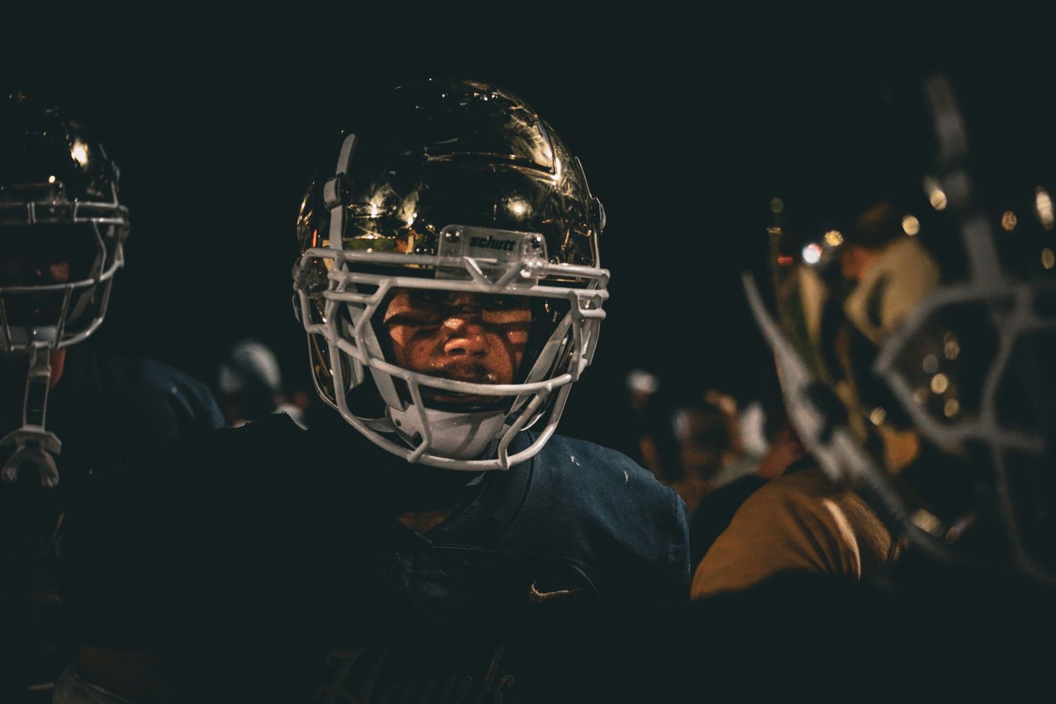 American football players in dark helmets and uniforms huddle together on the field during a night game.