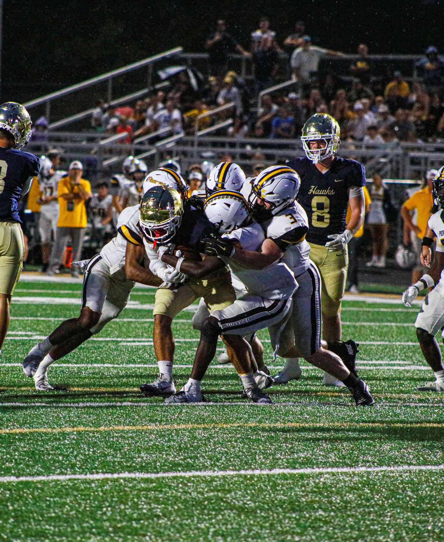 High school football players tackle ball carrier during intense night game on green turf field.