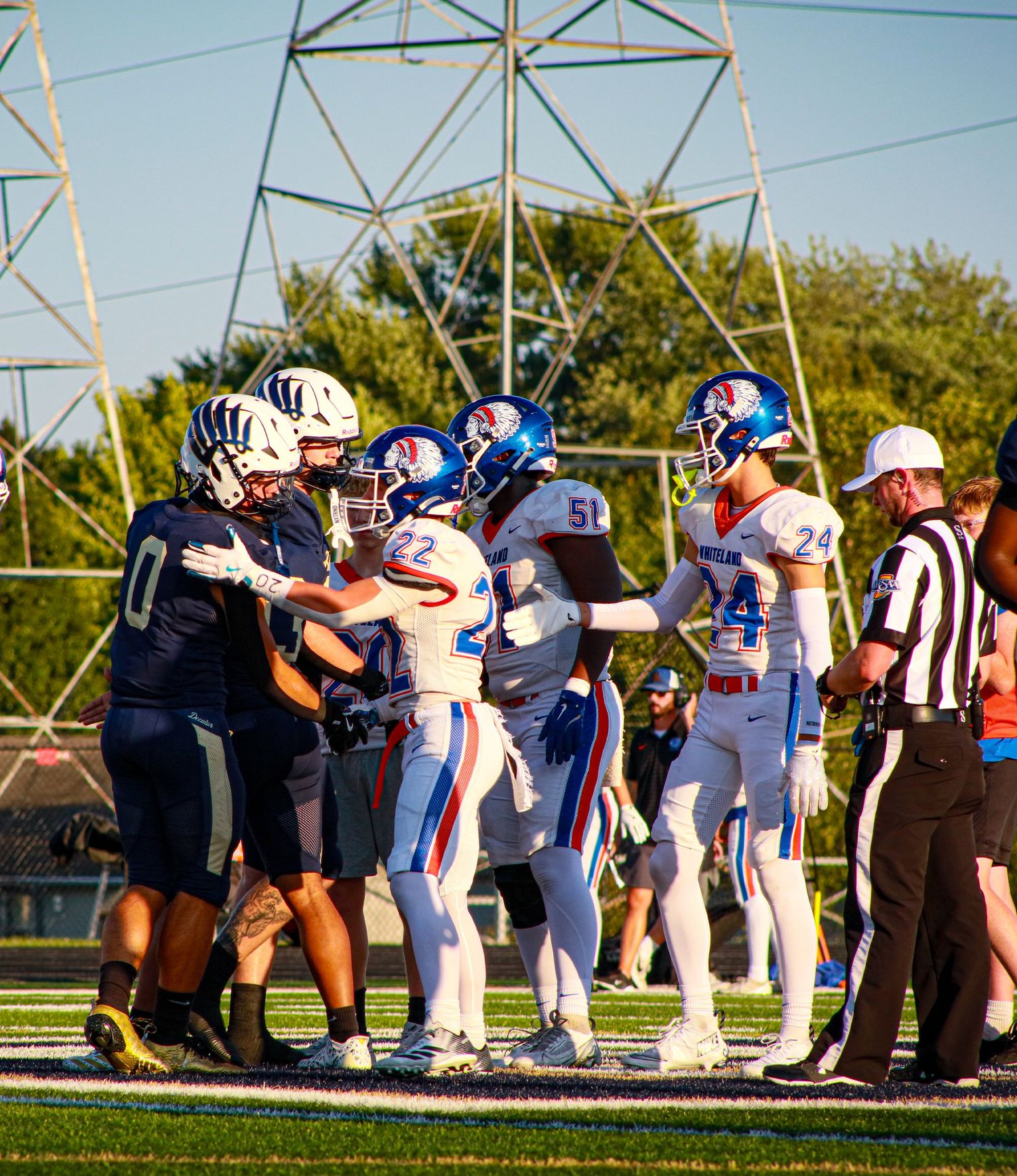 High school football players in blue and white uniforms face off against a dark jersey team during a game under stadium lights.