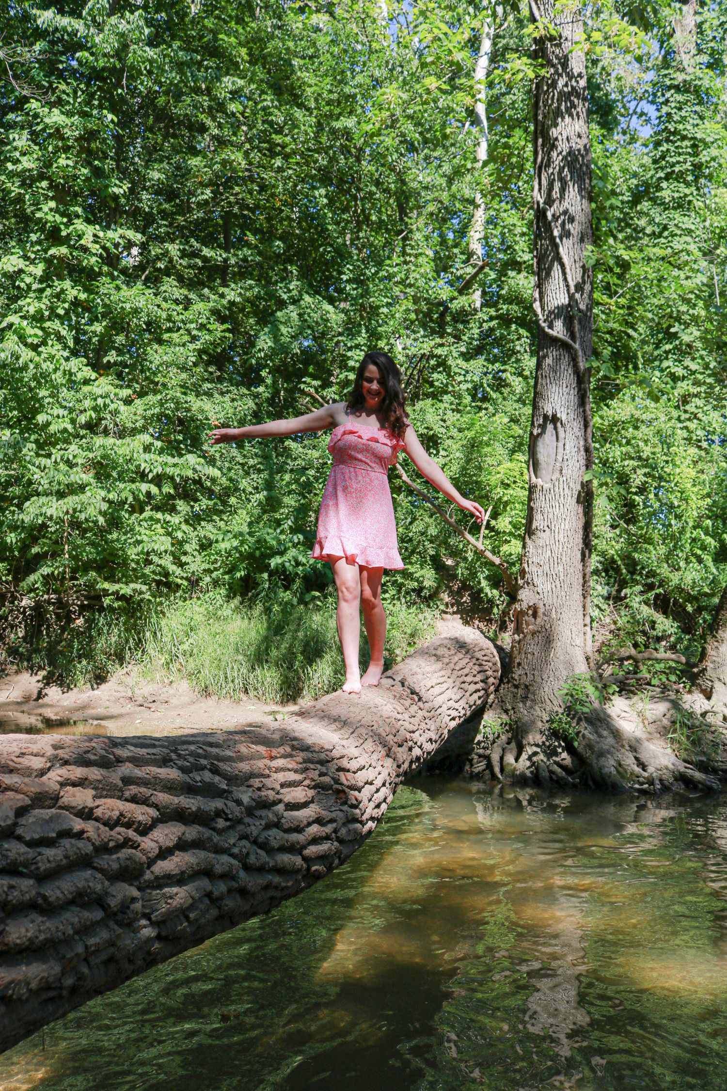 Woman in pink dress balancing on a fallen log over a stream in a lush green forest.