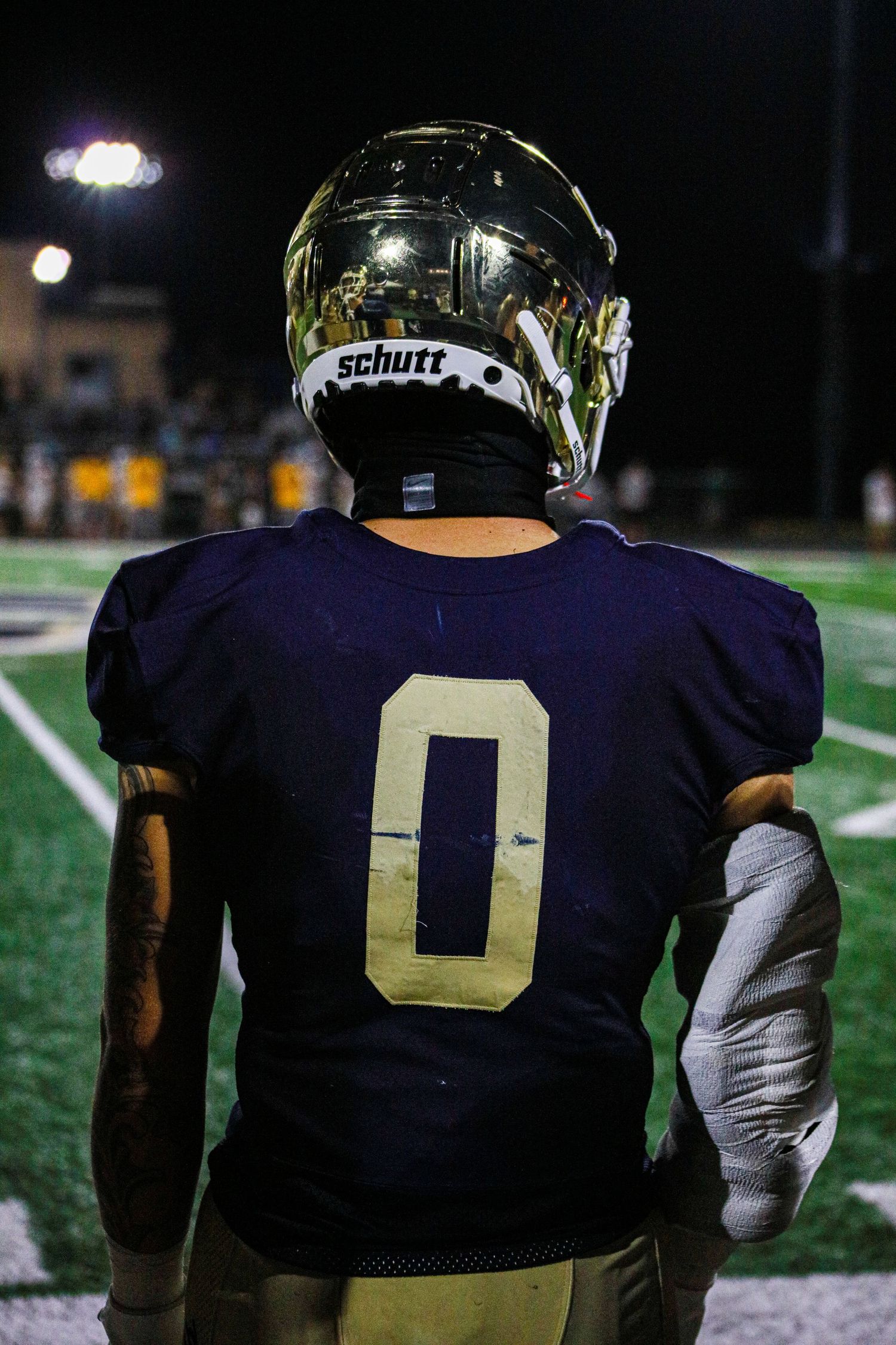 Football player wearing navy jersey number 0 and gold Schutt helmet stands on field during night game.