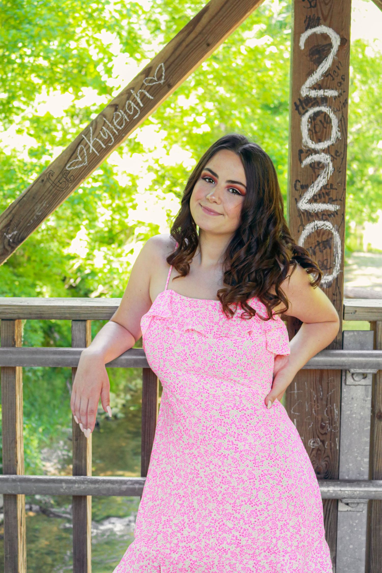 Young woman in pink floral sundress posing on wooden bridge with '2020' and 'Kayleigh' signs for senior photos.