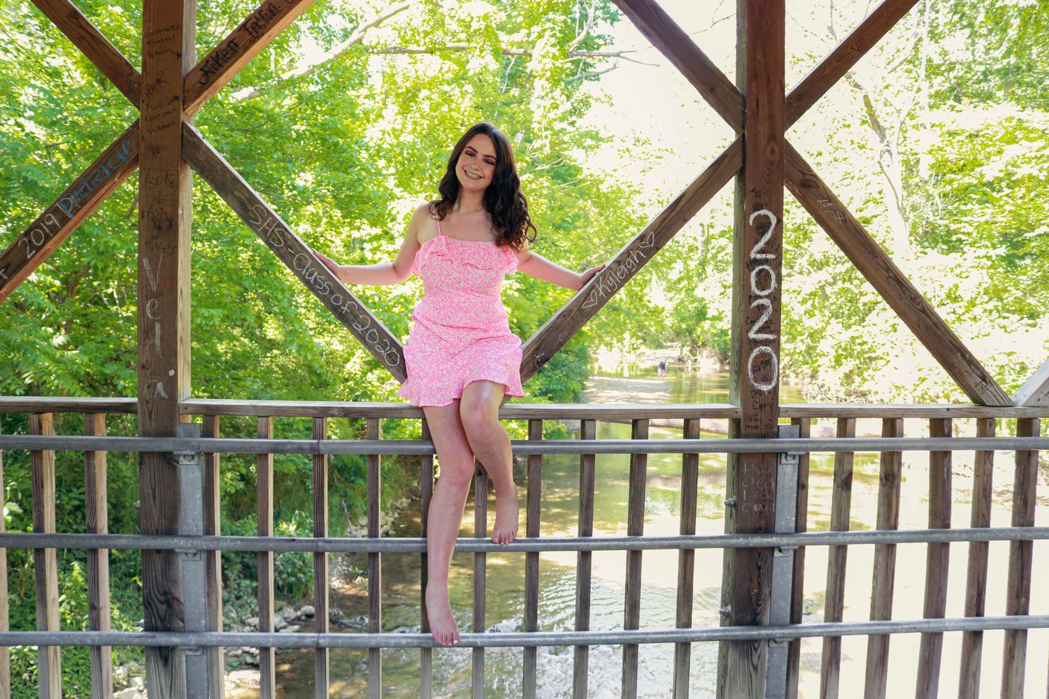 Young woman in pink dress posing on a wooden covered bridge surrounded by lush green trees, with 2020 sign.