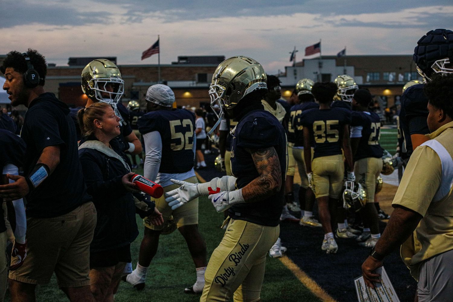 Football players in gold and navy uniforms gather on the sideline during an evening high school football game.