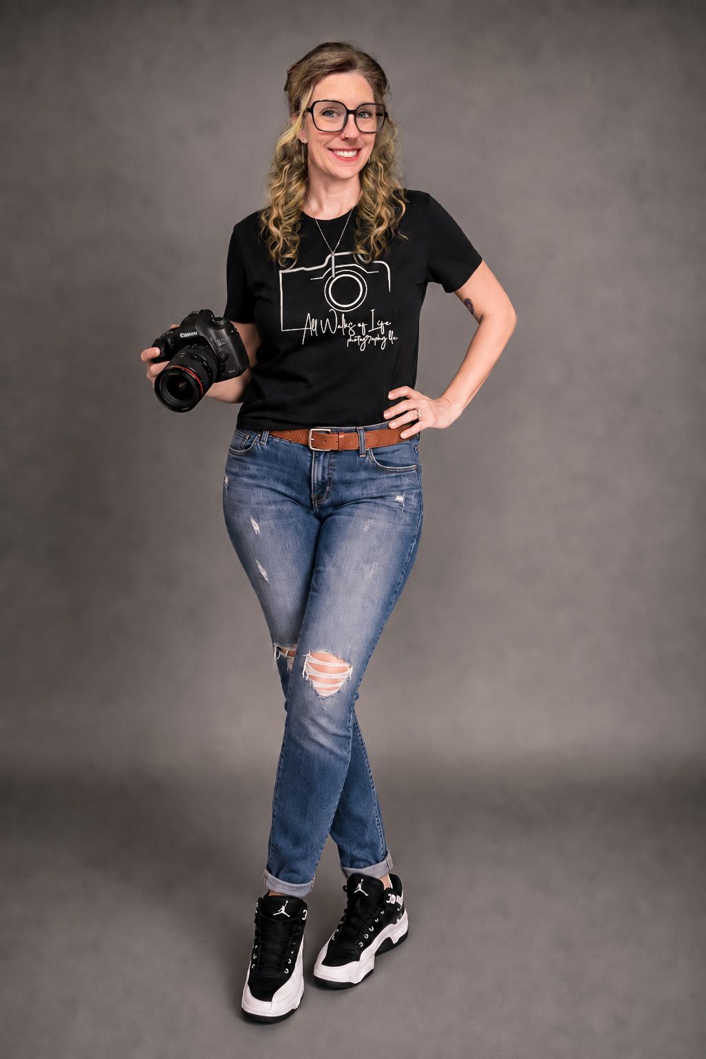 Smiling woman in black graphic tee, ripped jeans, and sneakers holding a DSLR camera against a gray background.