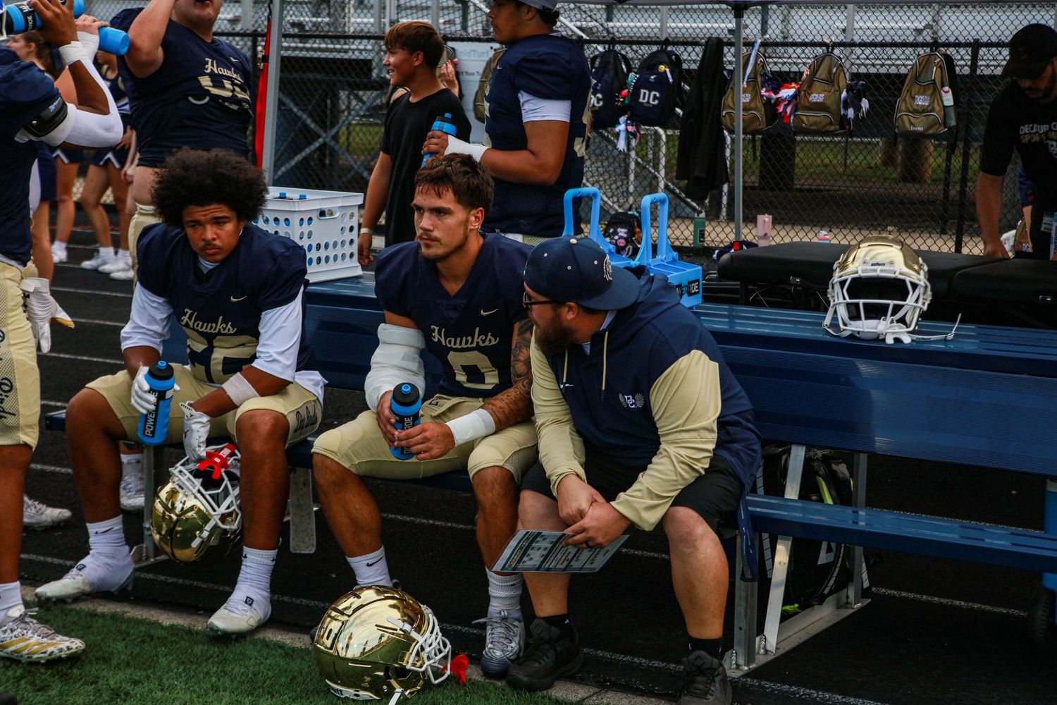 Football players in navy and gold uniforms rest on a bench during a game, holding helmets on a sunny day.