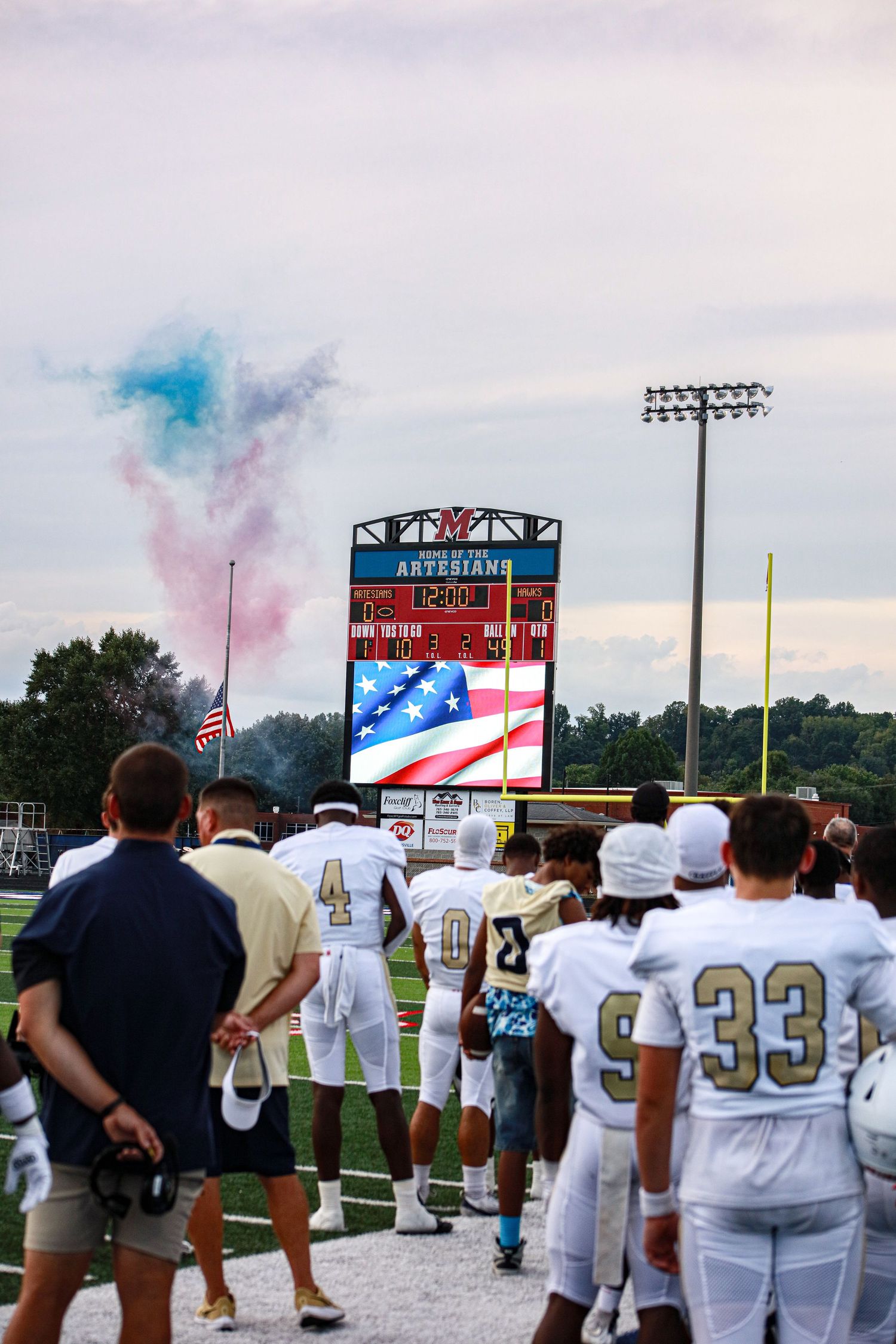 Football players stand at attention before a scoreboard displaying the American flag with colorful smoke during a pregame ceremony.