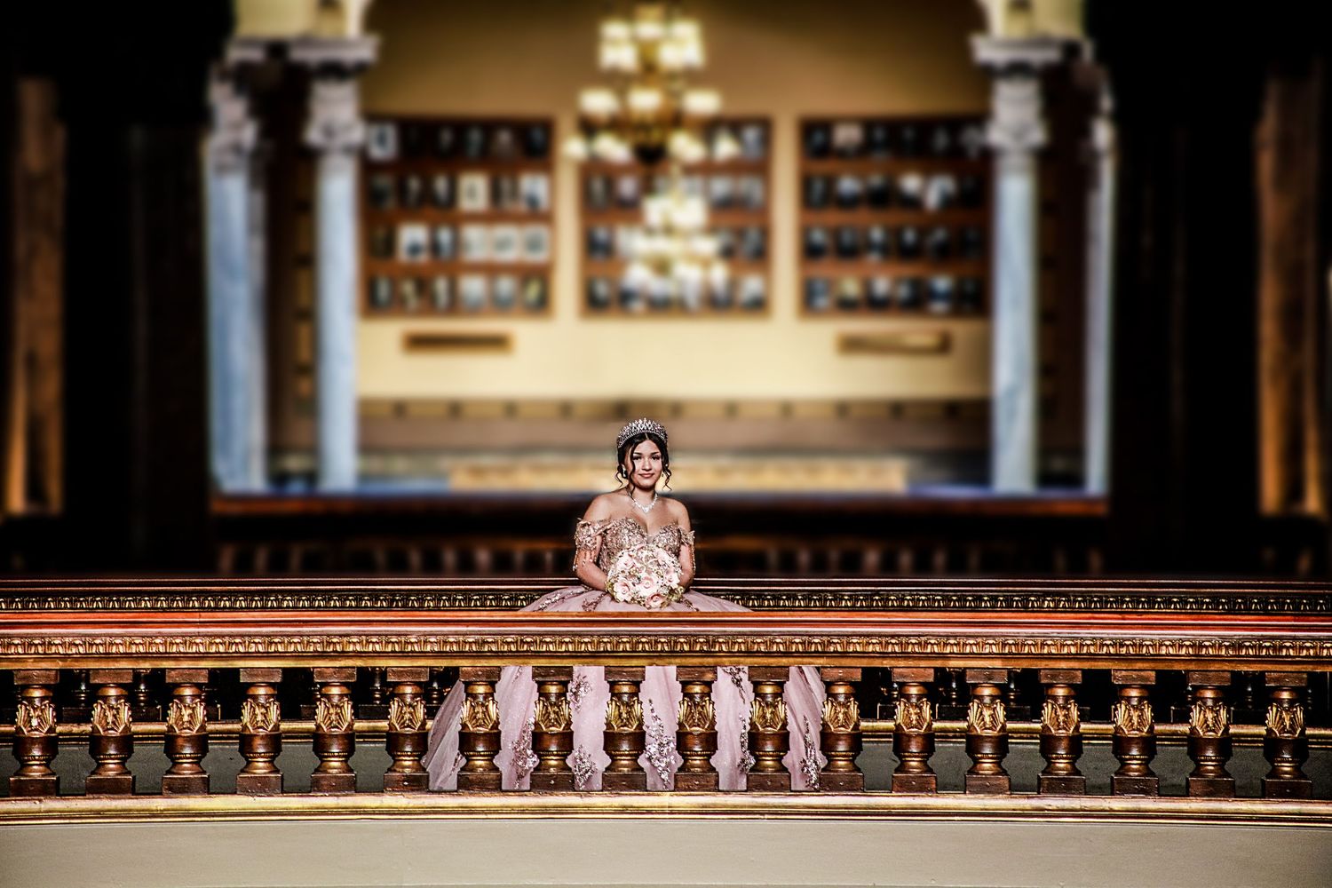 A woman in an elegant rose gold gown poses gracefully on a balcony inside a grand ornate building.