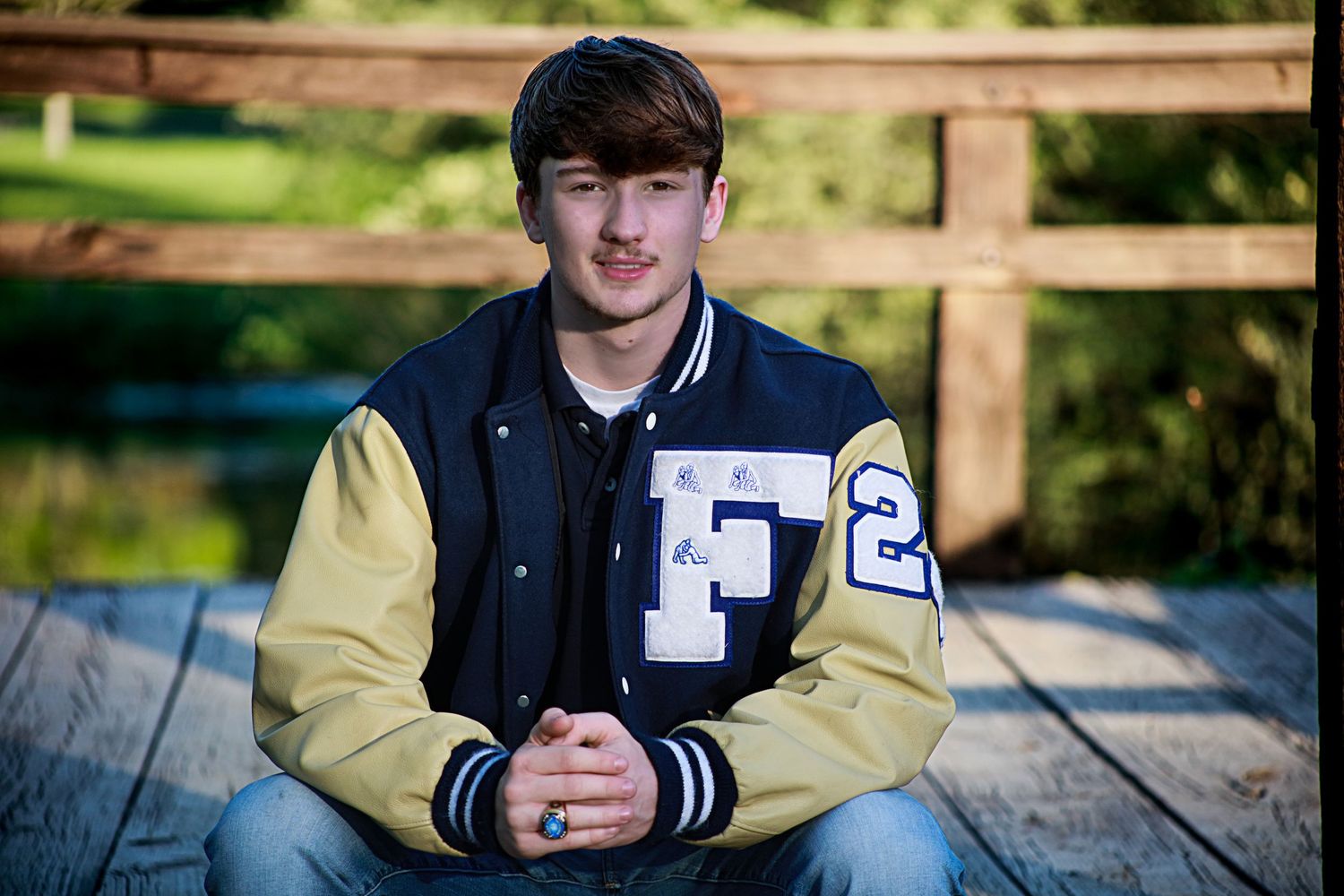 A teenage boy in a varsity letterman jacket sits on wooden steps near a bridge outdoors.