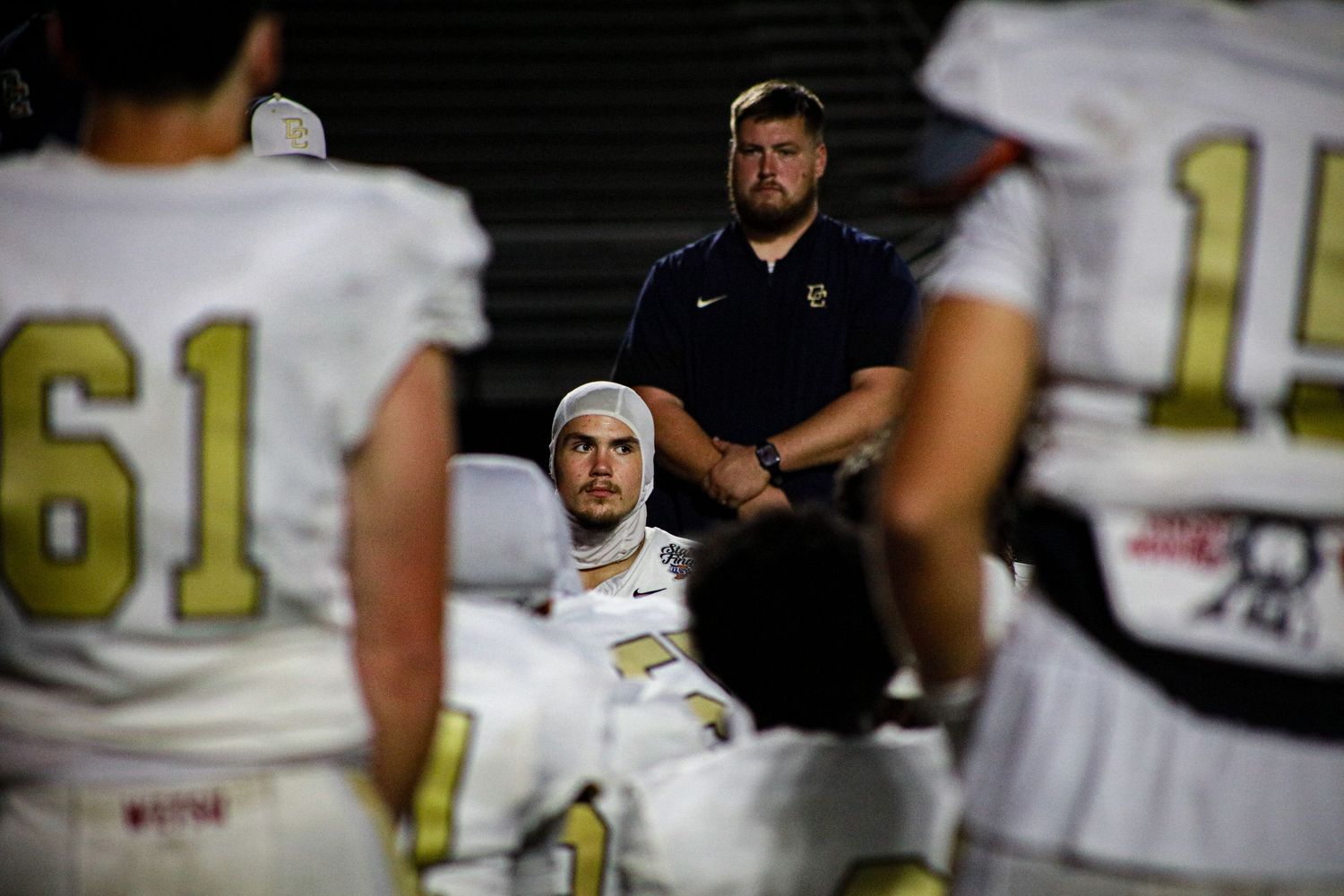 Football players in white and gold uniforms huddle around a coach during a nighttime game.