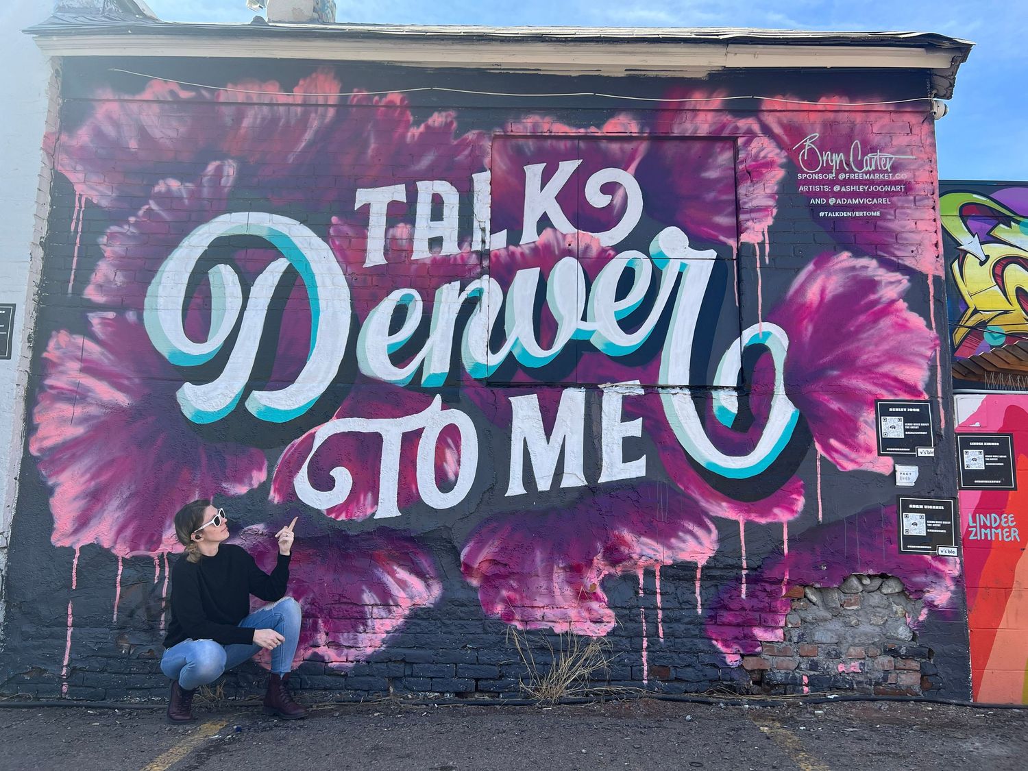 Person posing in front of a colorful Talk Denver To Me street mural with pink floral design in Denver, CO.