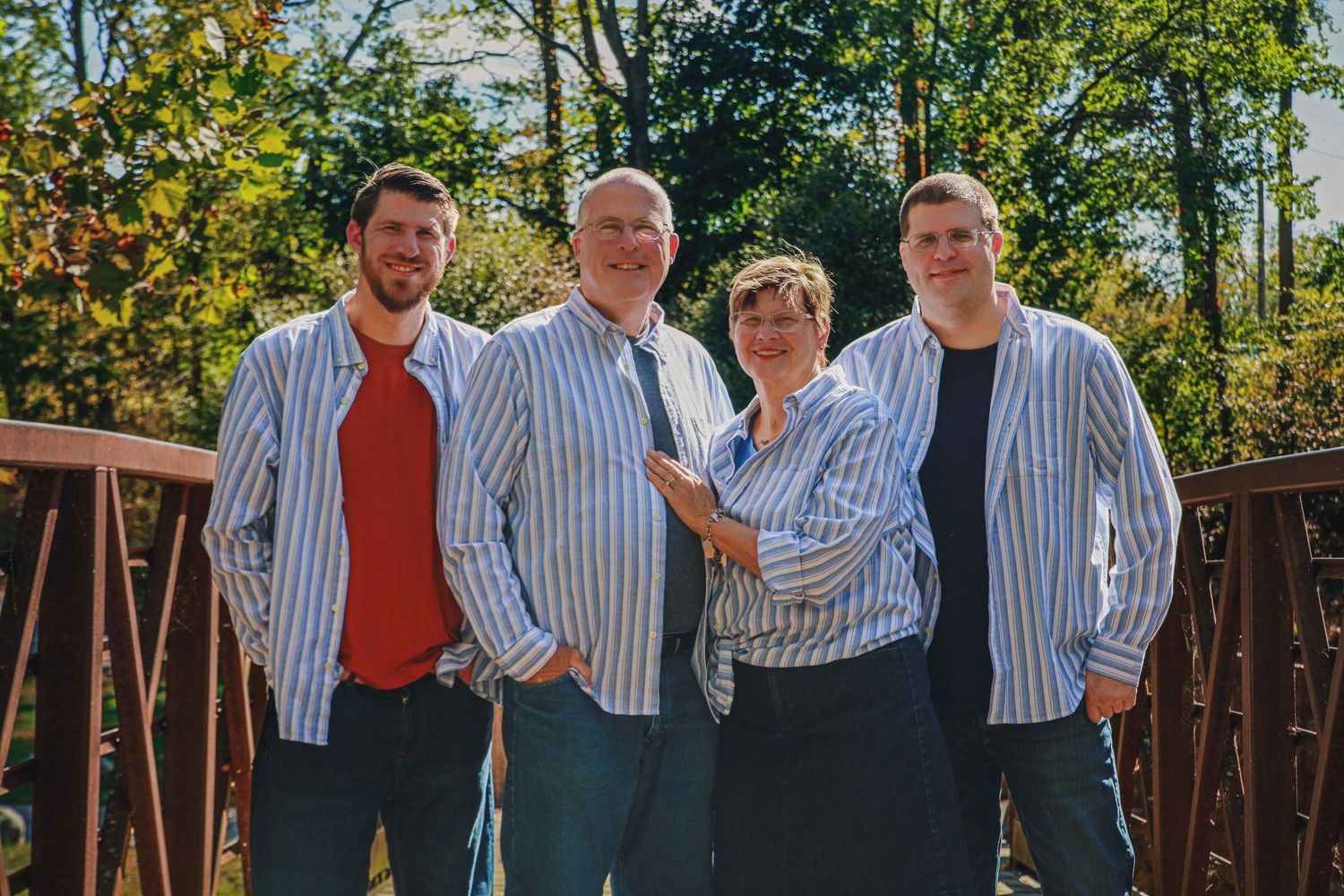 Family of four wearing matching blue striped shirts smiling together on a wooden bridge outdoors.