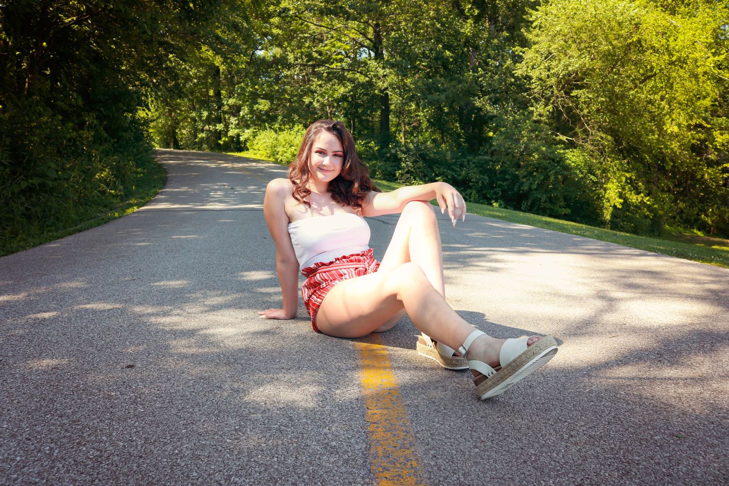 Young woman in red shorts sitting on a tree-lined road, smiling confidently in casual summer attire.