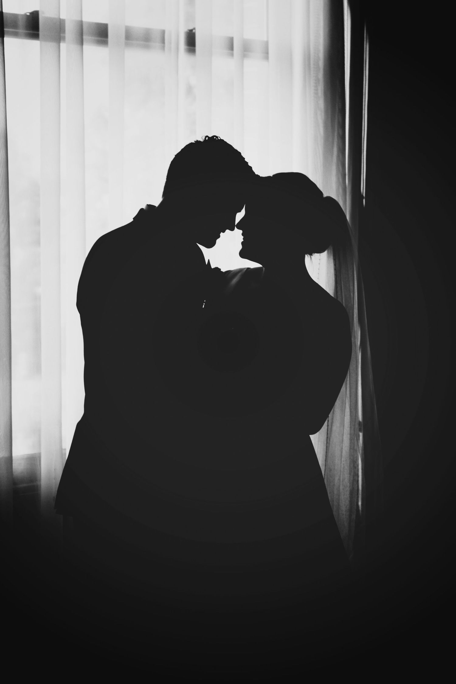 Black and white silhouette of a romantic couple standing close together in front of a bright window with sheer curtains.