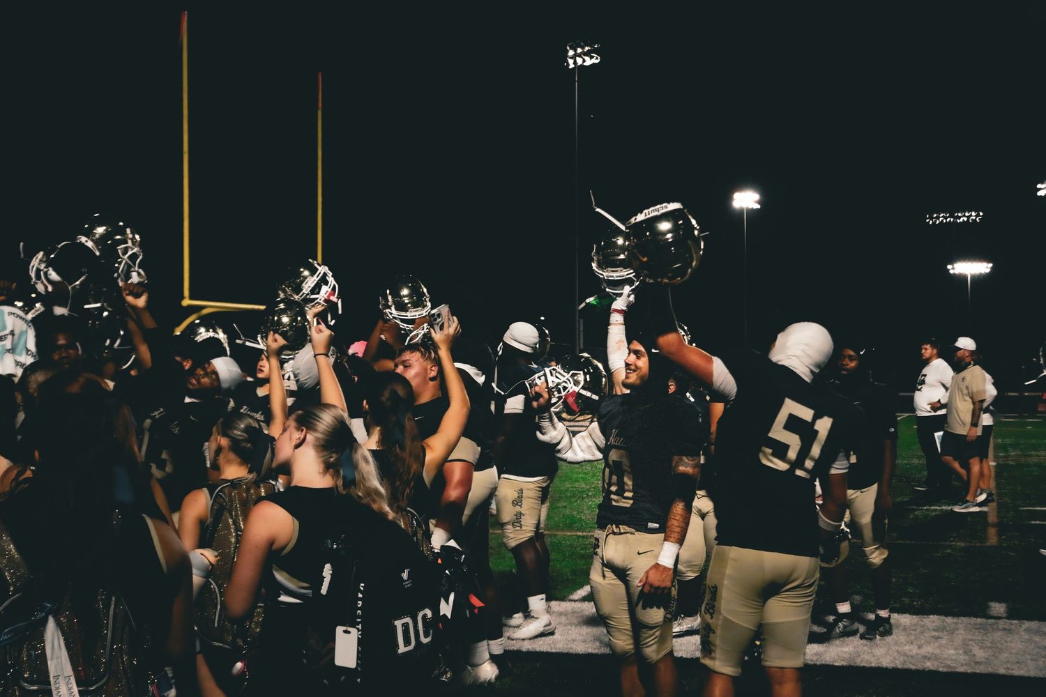 Football players and fans celebrate on field at night, player #51 raises helmet in victory under stadium lights.
