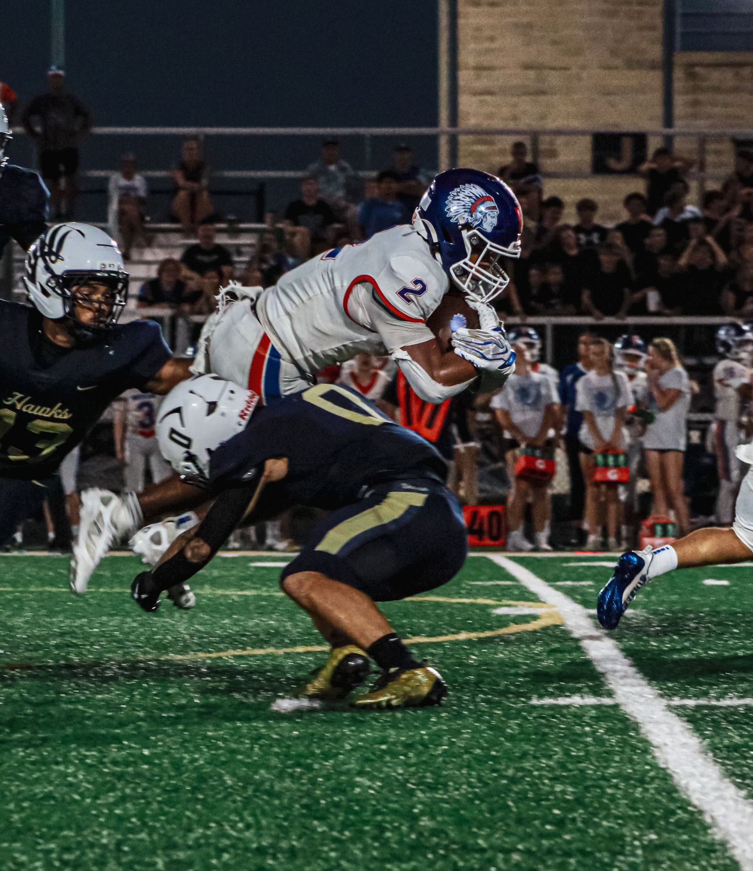 High school football player in white uniform leaping over defender during night game on green turf field.