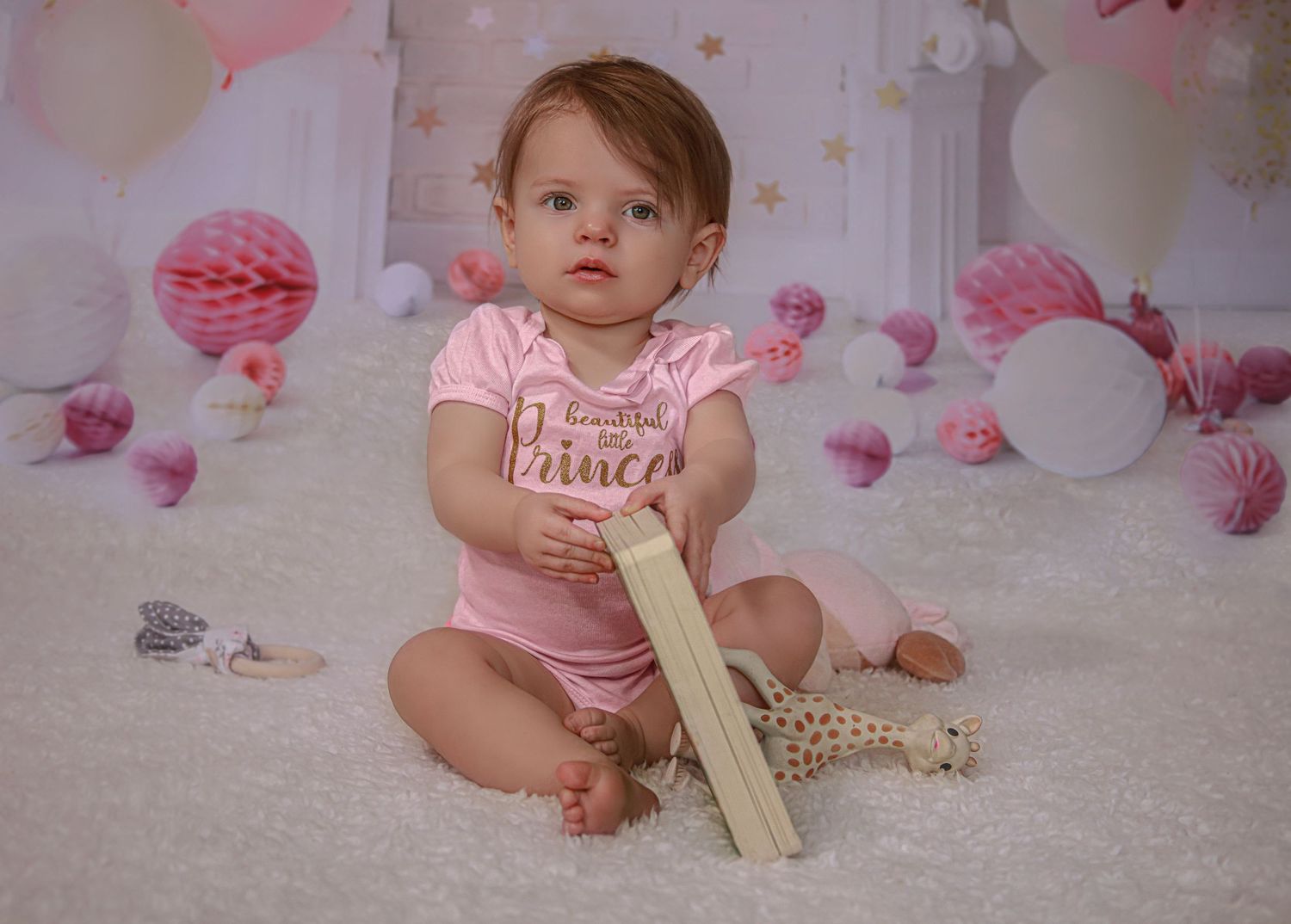 Curious baby girl in pink princess onesie holds wooden toy while sitting on white fluffy rug.