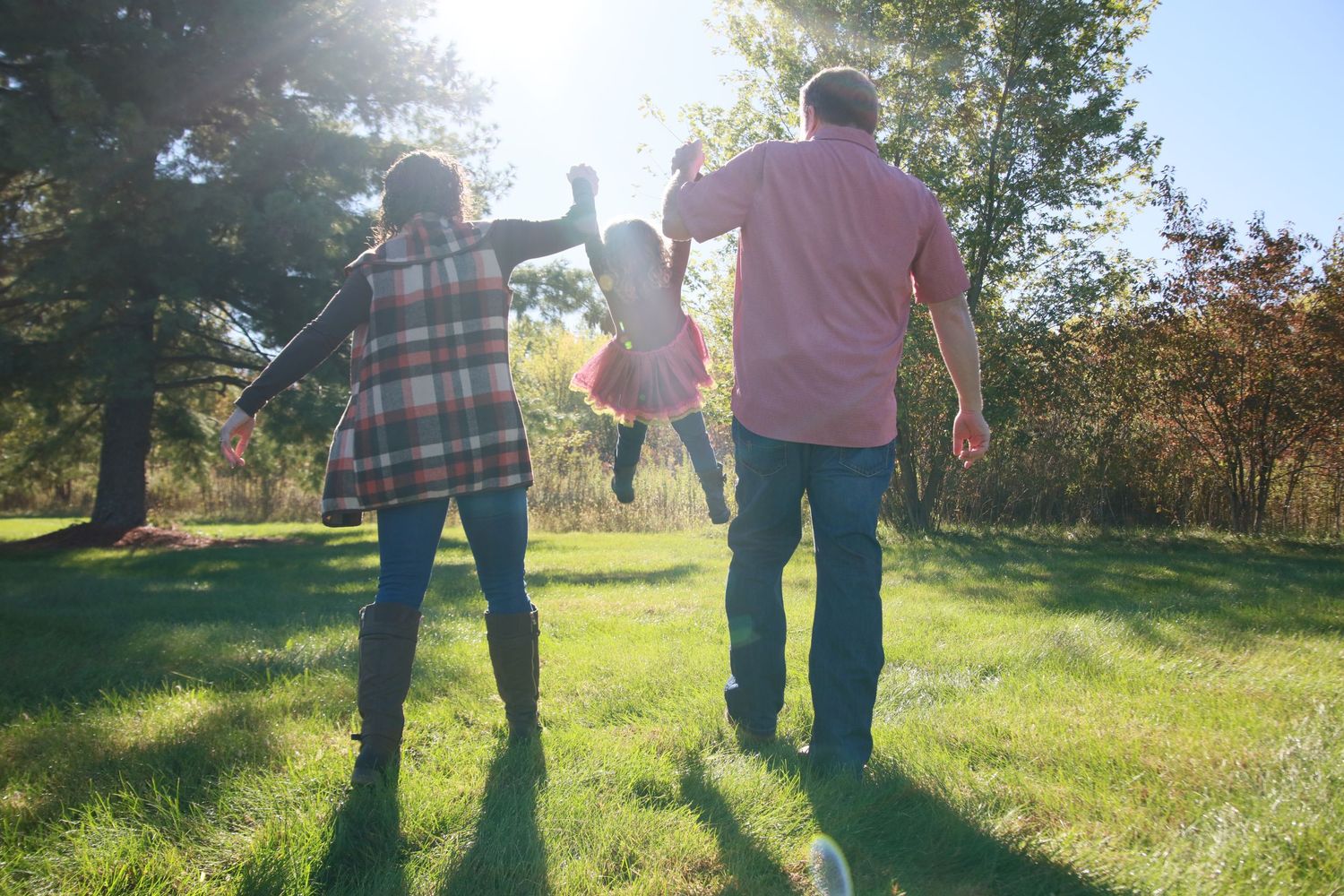 Family of three holding hands walking through sunny green park in autumn.