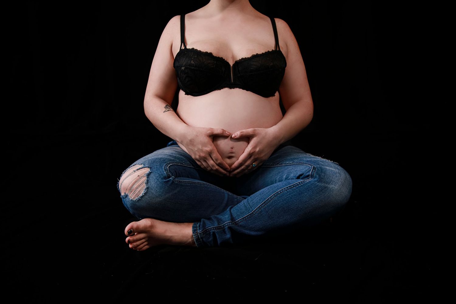 Woman sitting on the floor with pregnant belly out and hands in the shape of a heart laying gently on belly.