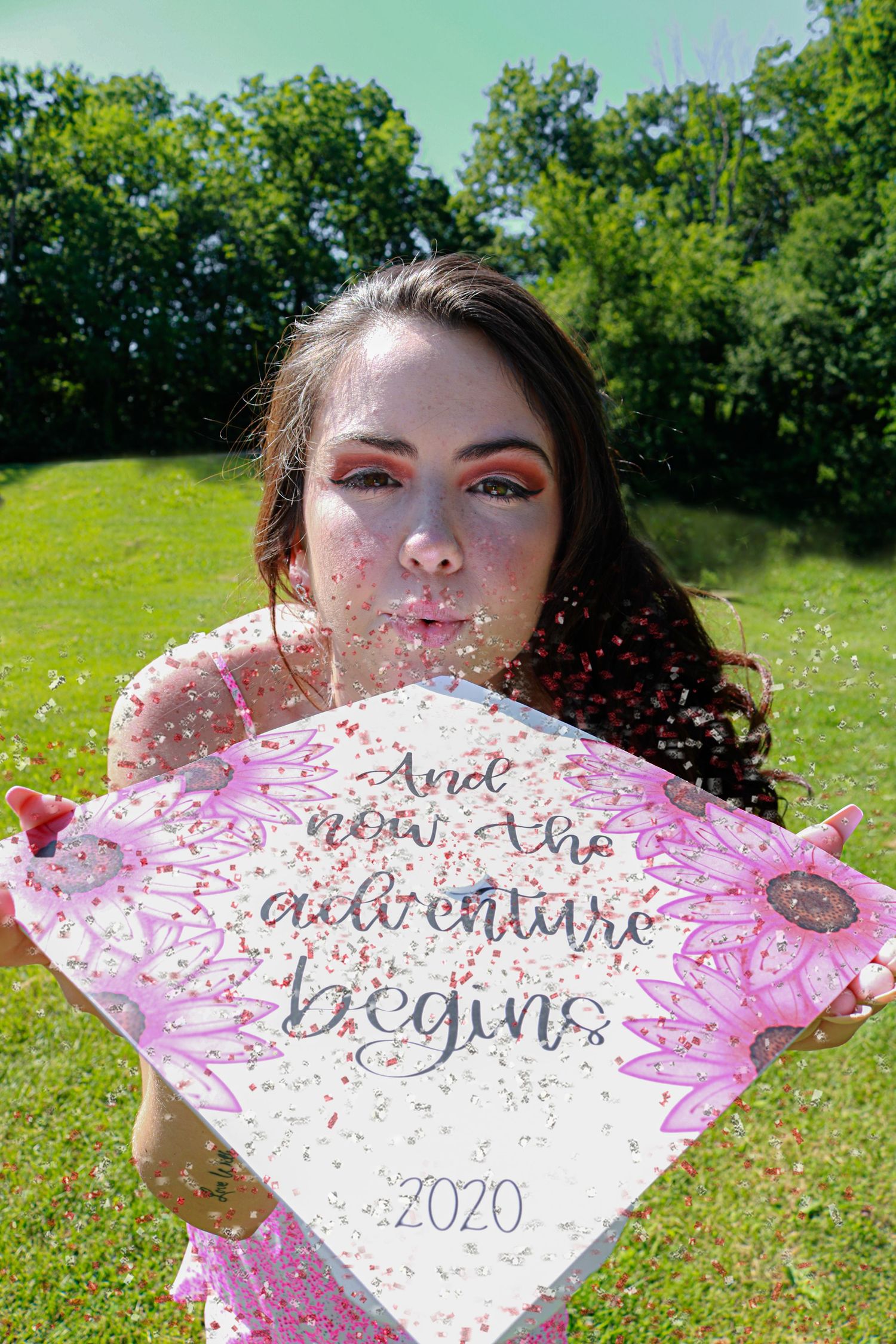 Graduate blowing confetti while holding decorated 2020 cap reading 'And now the adventure begins' outdoors.