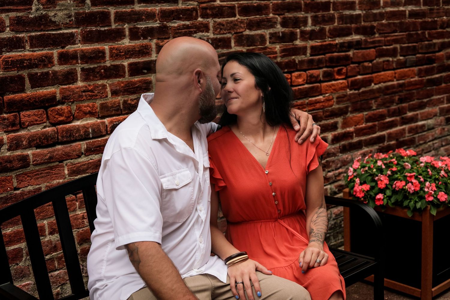 Bald man and dark-haired woman in red dress sharing romantic moment by brick wall.