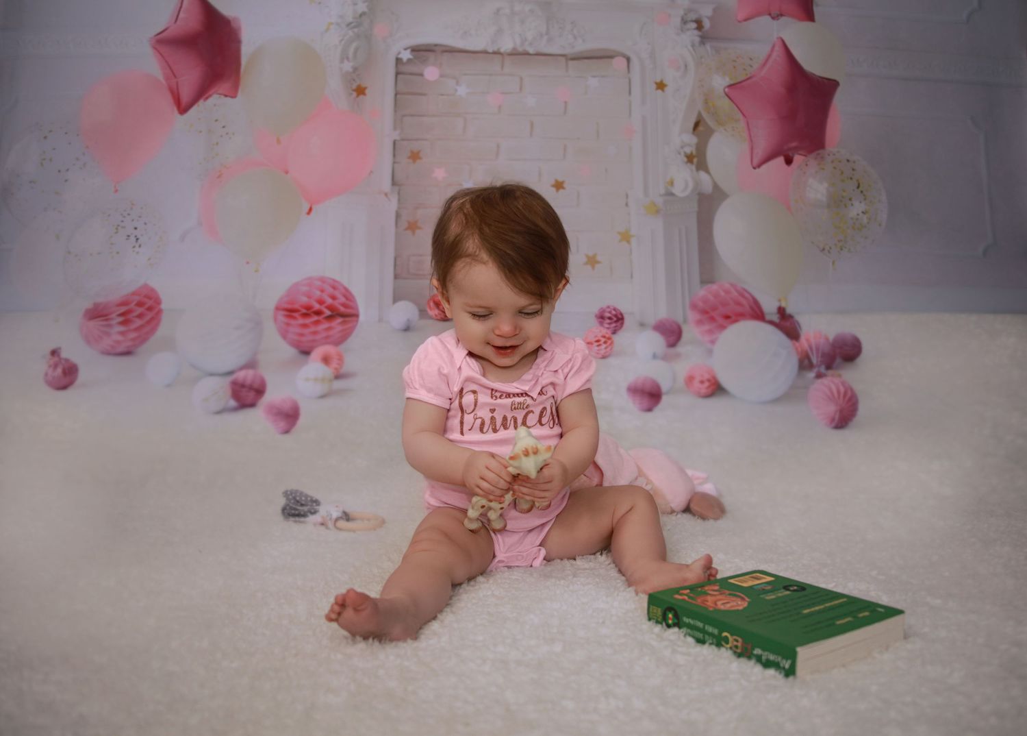 Smiling baby girl in pink outfit plays with book during first birthday cake smash photo session.