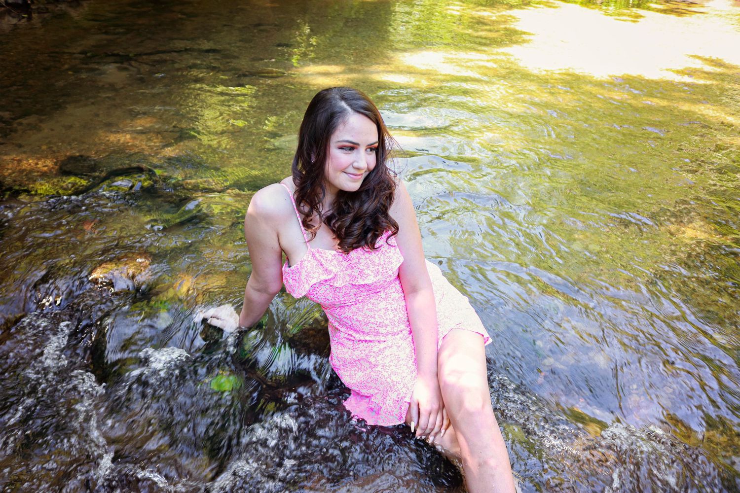 Young woman in pink floral dress sitting on rocks in a shallow sunlit stream.
