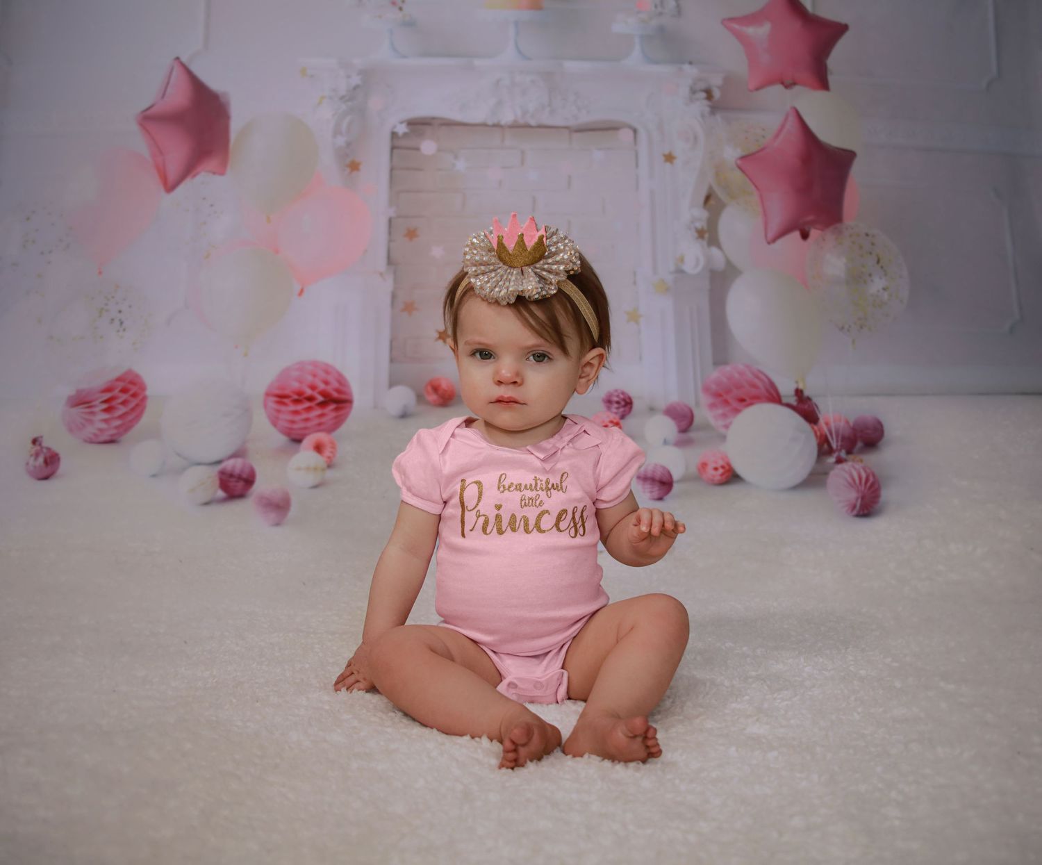 Baby girl in pink princess onesie with floral headband sits before pink balloon birthday backdrop.