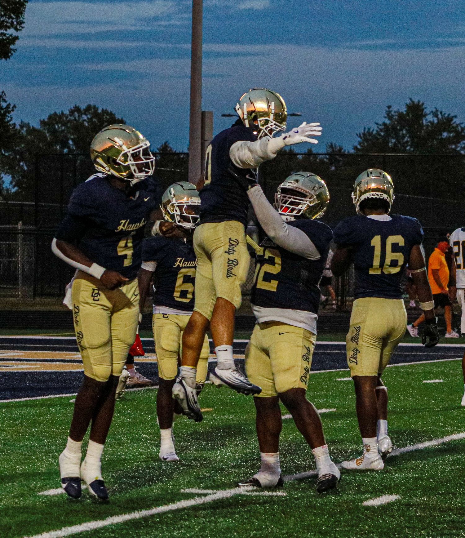 High school football players in navy and gold uniforms celebrate a touchdown on the field during an evening game.